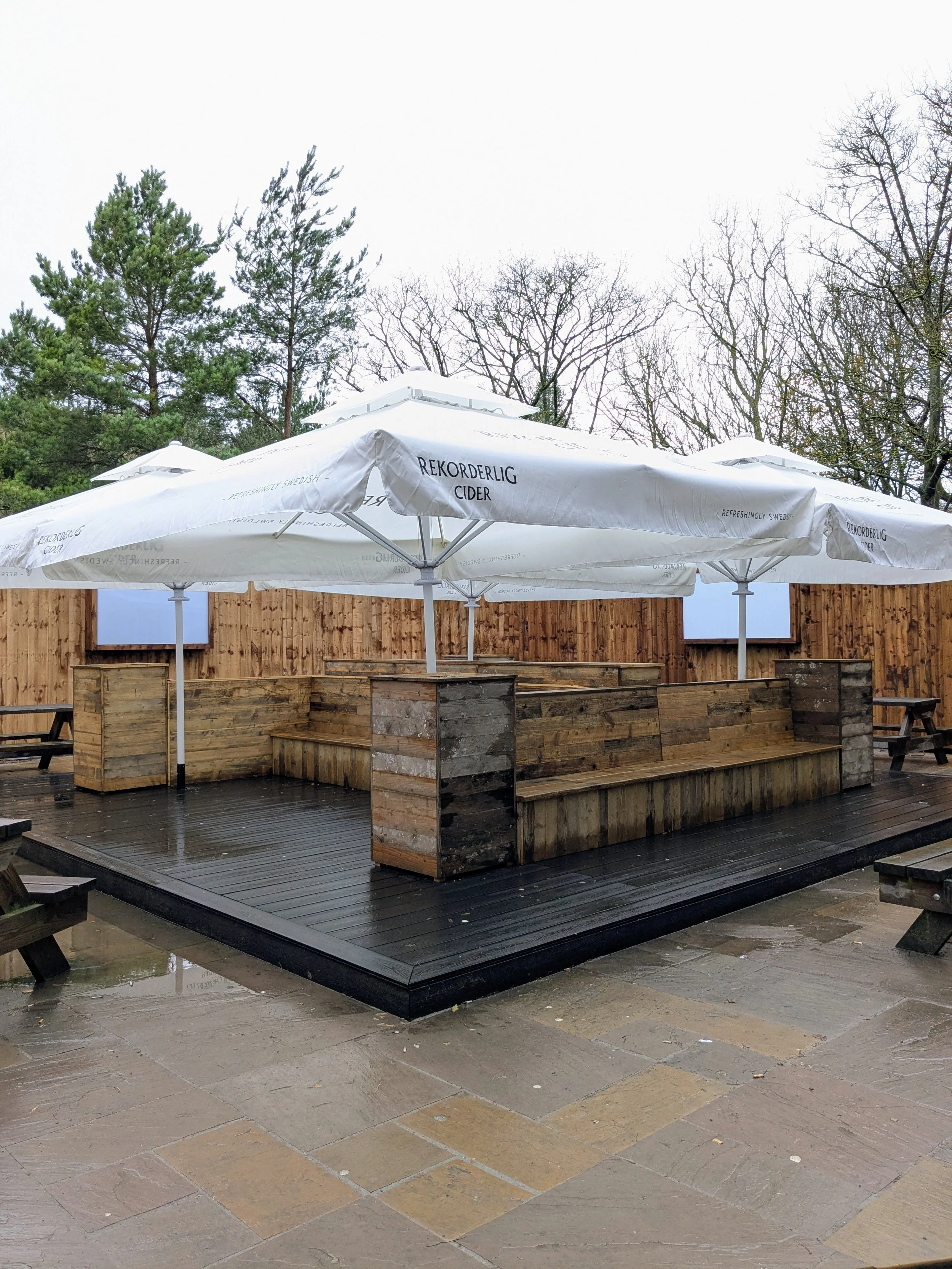 Empty outdoor patio with wooden seating area, large white umbrellas labeled 'Rekorderlig Cider,' and a wooden fence, with trees in the background.