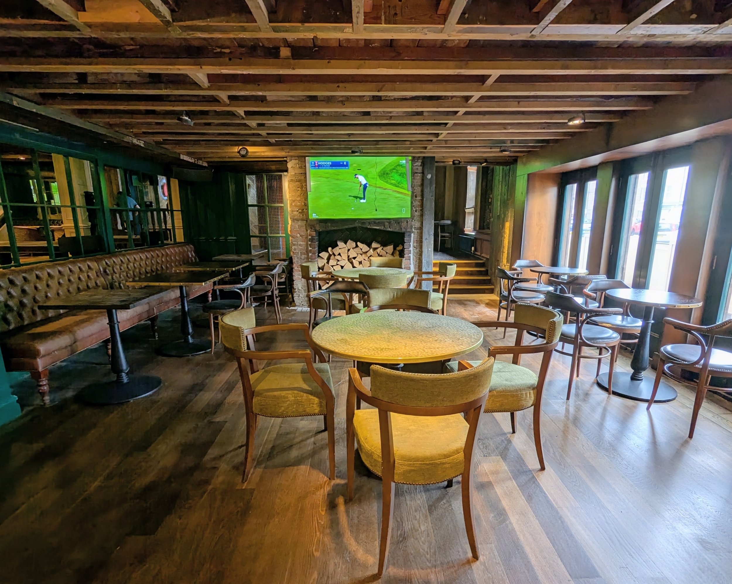 Interior view of a restaurant or pub with wooden ceiling beams, a fireplace with firewood stacked inside, a television showing a golf game, and various chairs and tables.