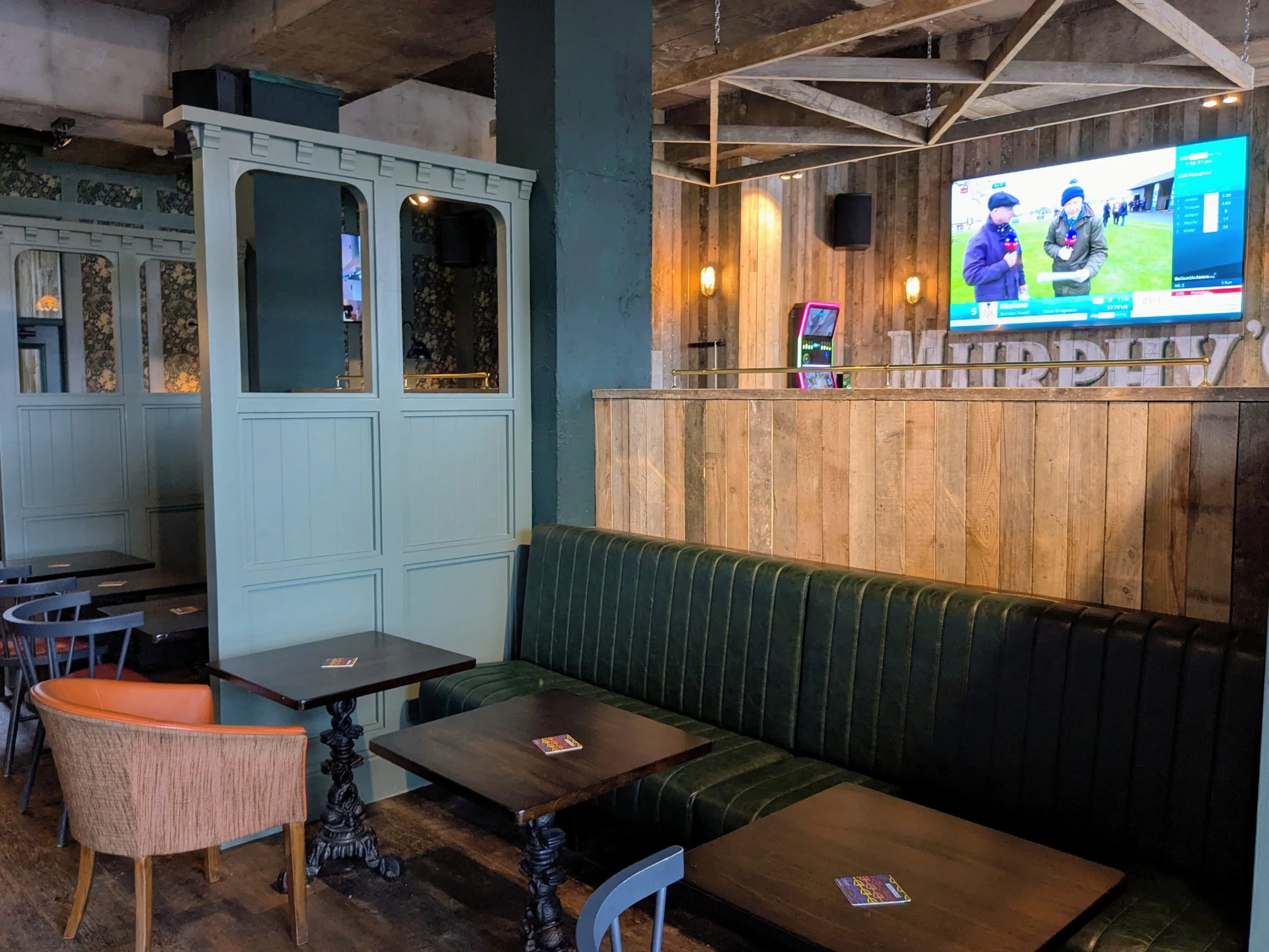 Interior of a restaurant or bar with wooden accents, a large TV on the wall showing a race, and various chairs and tables.
