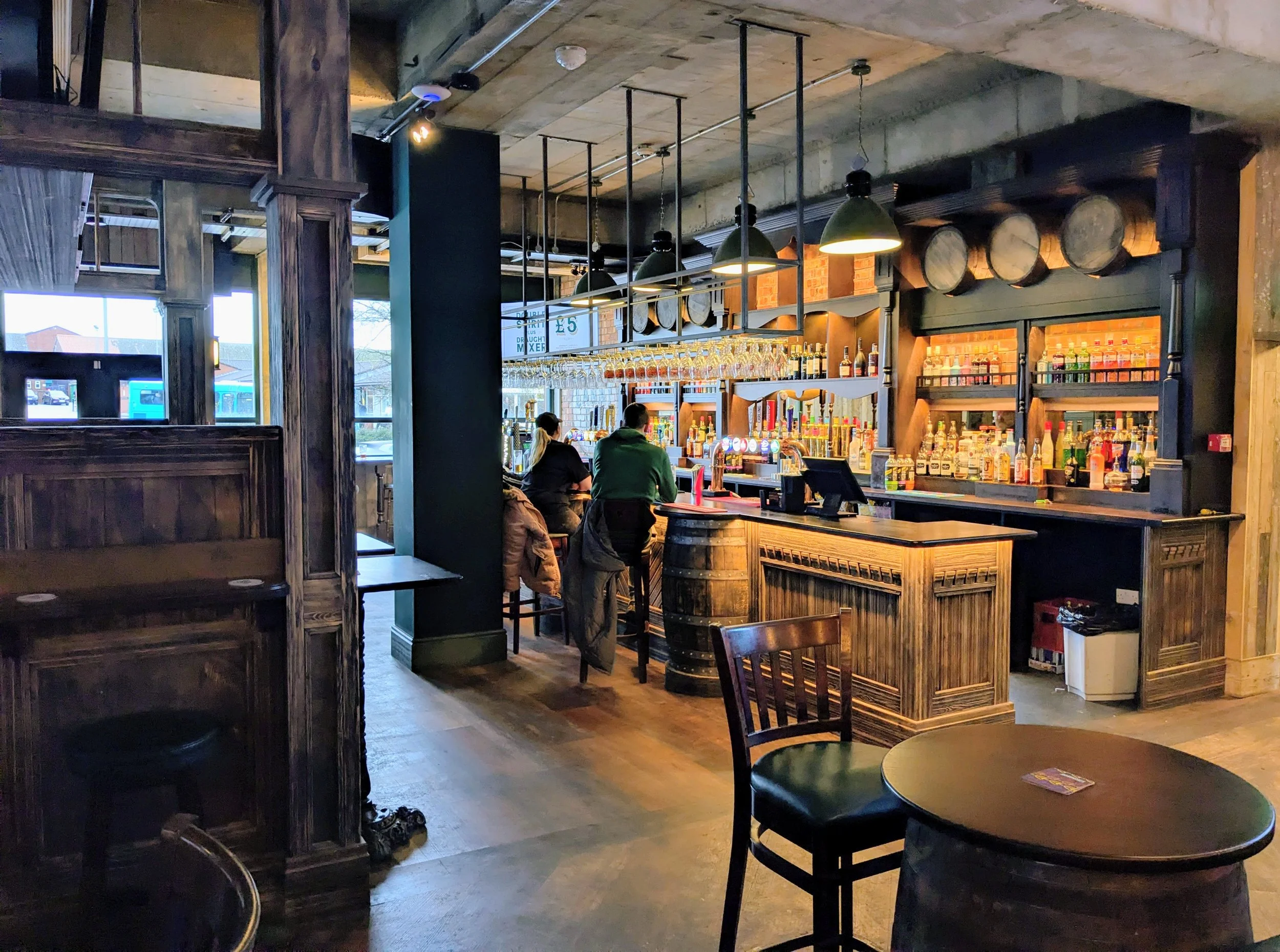 Inside a rustic bar with a wooden bar counter, hanging wine glasses, and barrels on the wall. Two people are seated at the bar. There are tables and chairs in the foreground, with natural light coming through the windows.