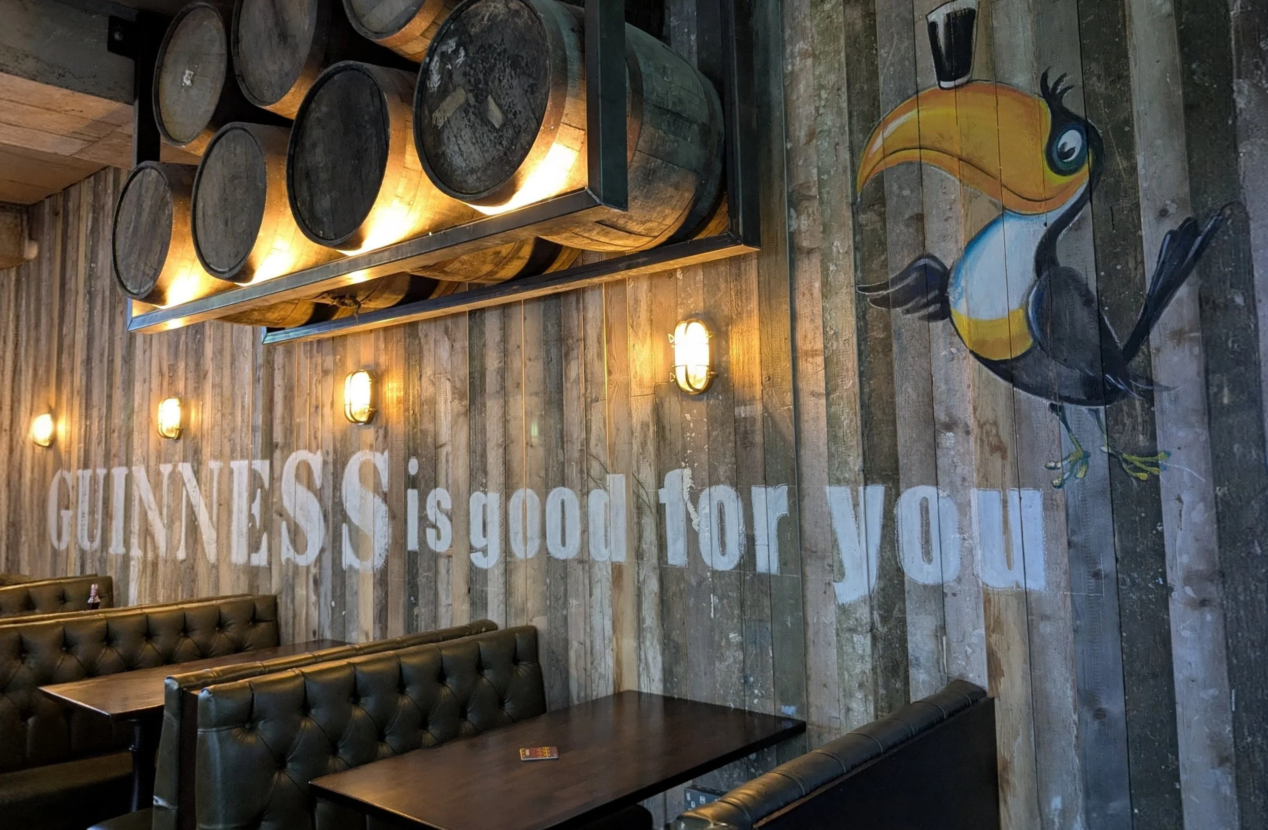 Interior of a rustic restaurant or bar with wood-paneled walls, leather booths, and a wooden table. A mural of a colorful toucan painted on the floor, and a display of wine barrels on a metal rack illuminated by warm lighting. The wall features large, white text.