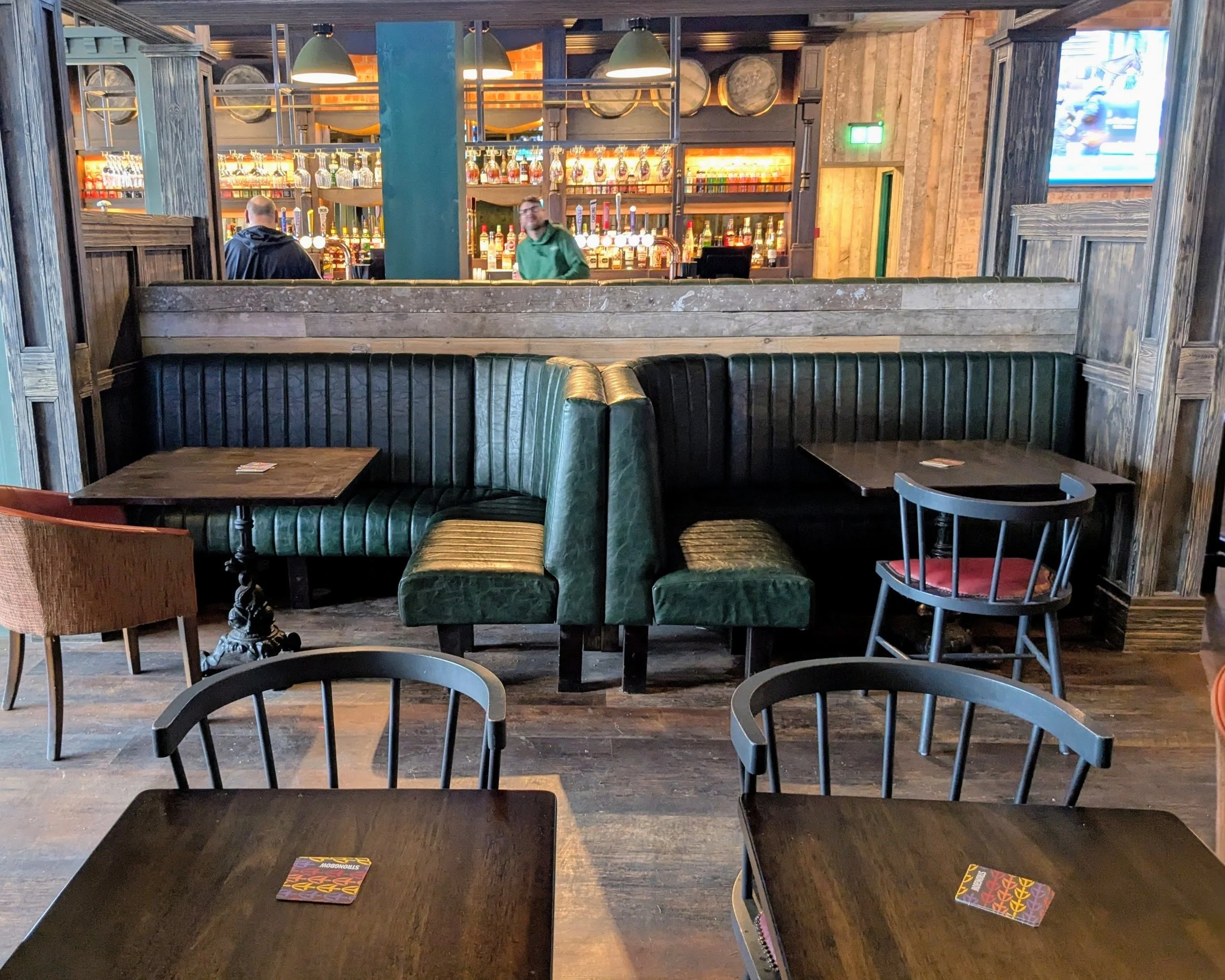 Interior of a bar or pub with tables, chairs, and a bar counter in the background. There are two people standing near the bar, and a large television screen on the wall displaying a sports game.