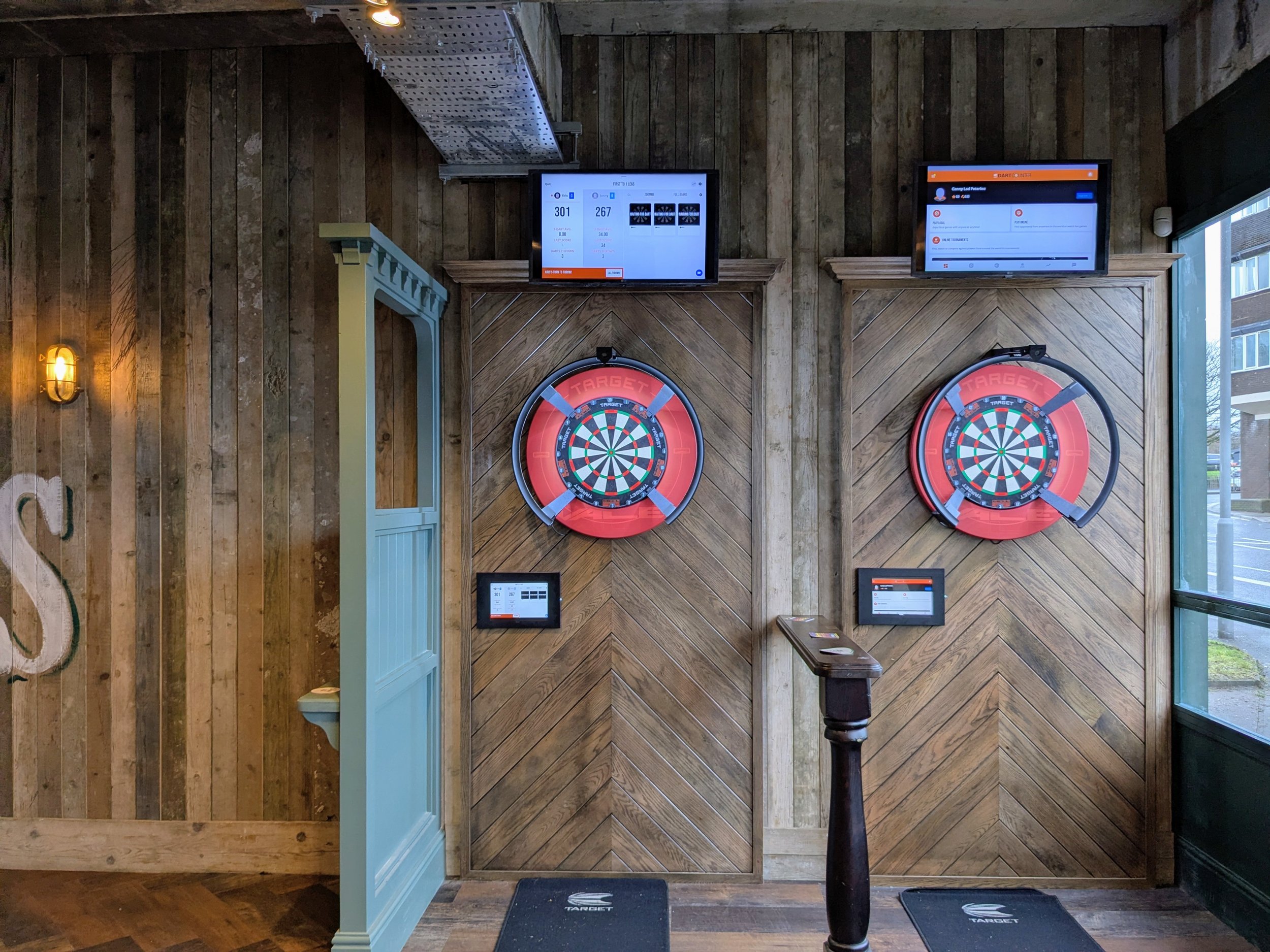 Indoor darts area with two electronic dartboards mounted on a wood-paneled wall, each with a targeting area below, and two screens displaying scores and game information mounted above. A small table and a standing monitor are positioned near the dartboards.