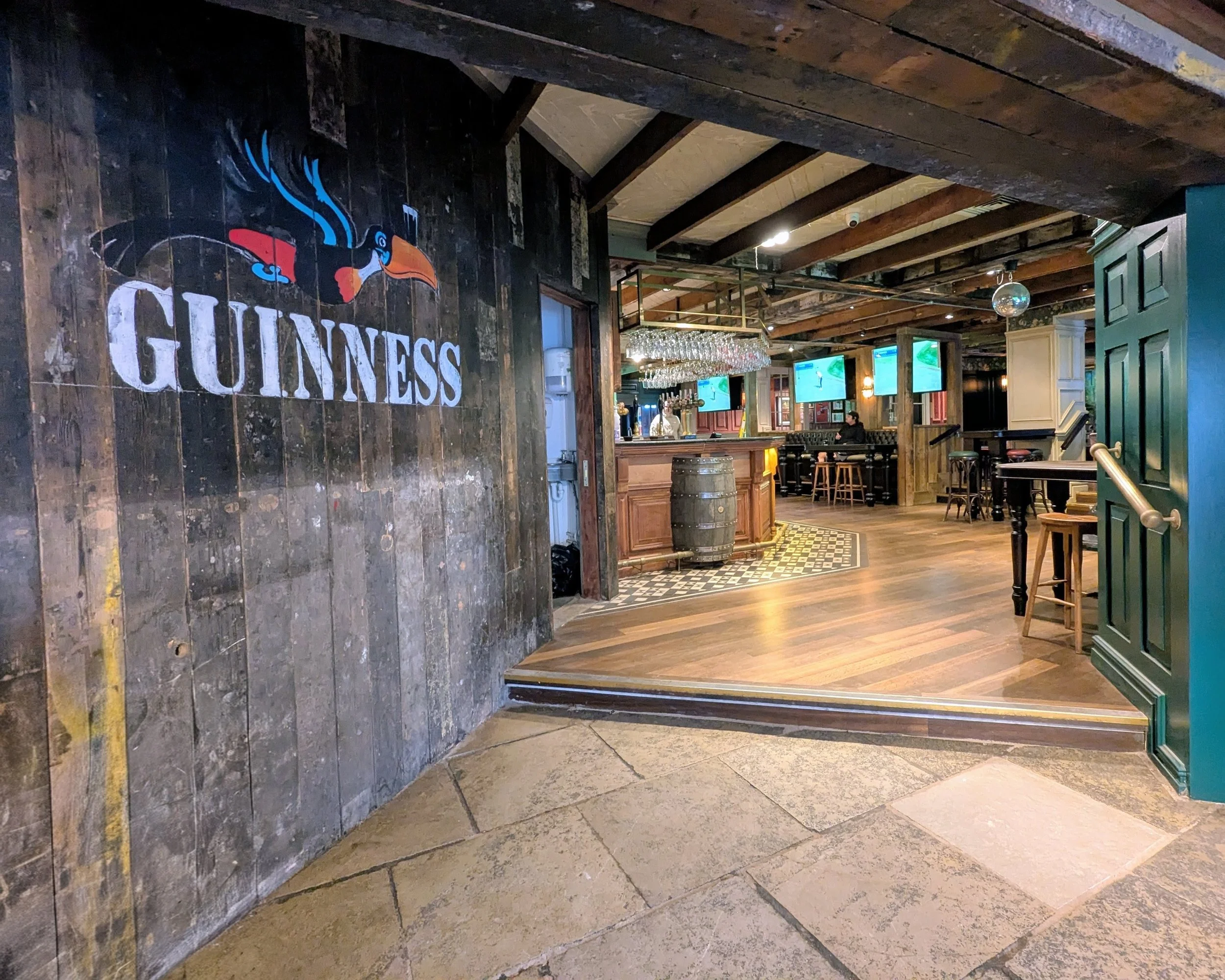 An interior of a bar or pub with wooden ceiling beams, a wall with a Guinness logo featuring a toucan, a mirror, multiple television screens, a bar counter, barstools, and a person sitting at the bar.