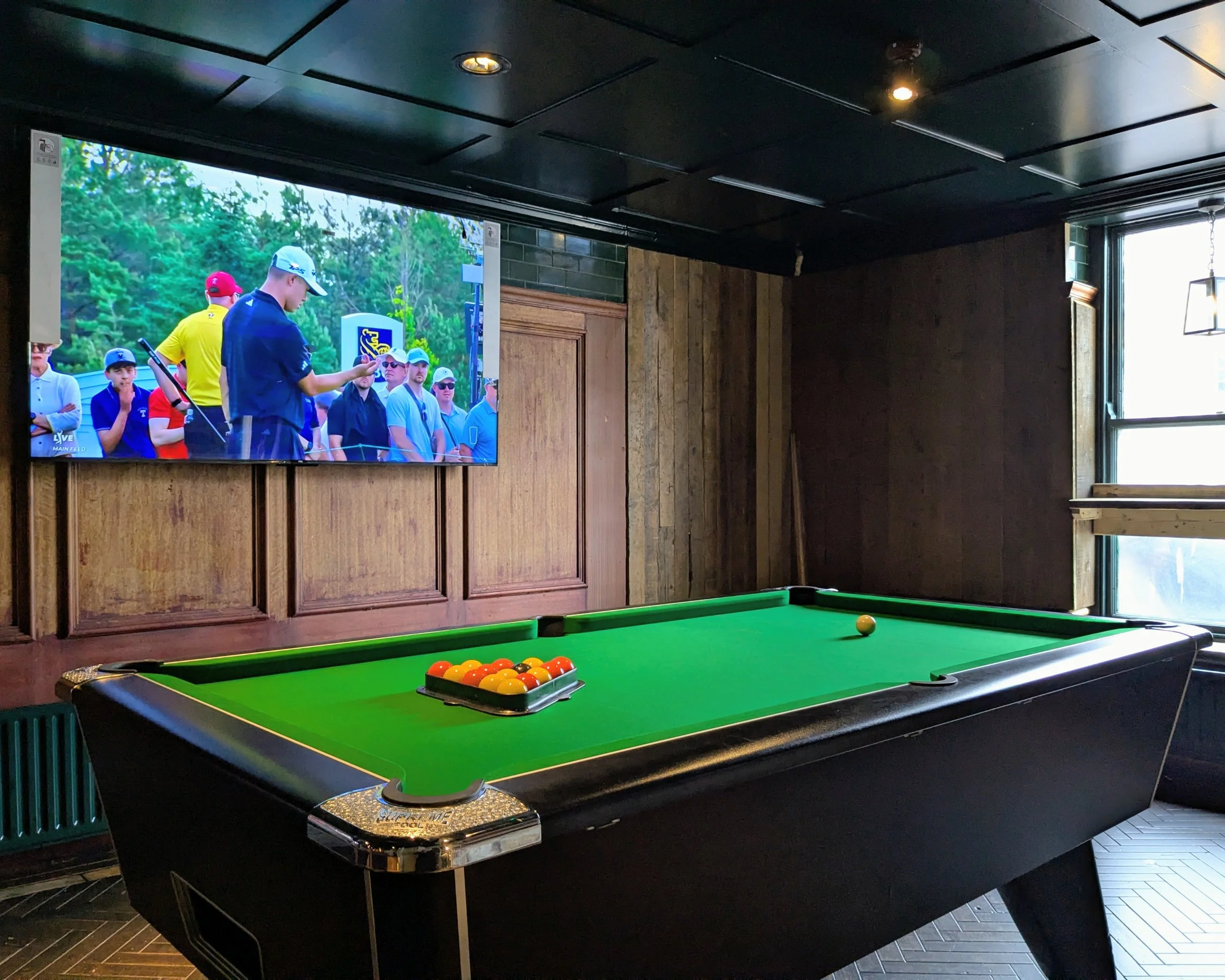 Indoor billiards table with green felt and pool balls, in a room with dark walls, wood paneling, and a large window.