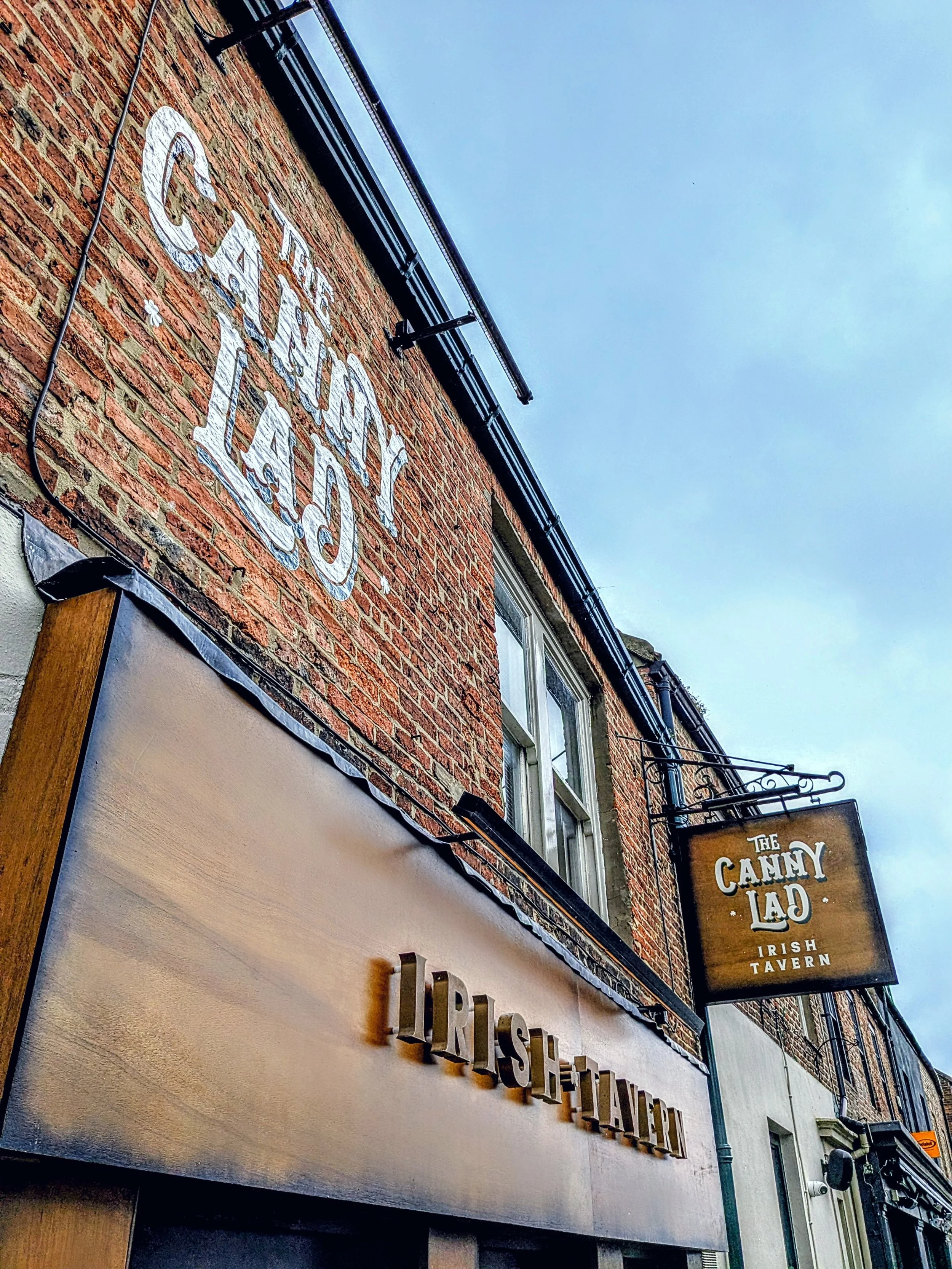 Exterior view of a brick building with signs for The Canny Lad Irish Tavern and a restaurant, with windows and a black metal balcony, under an overcast sky.