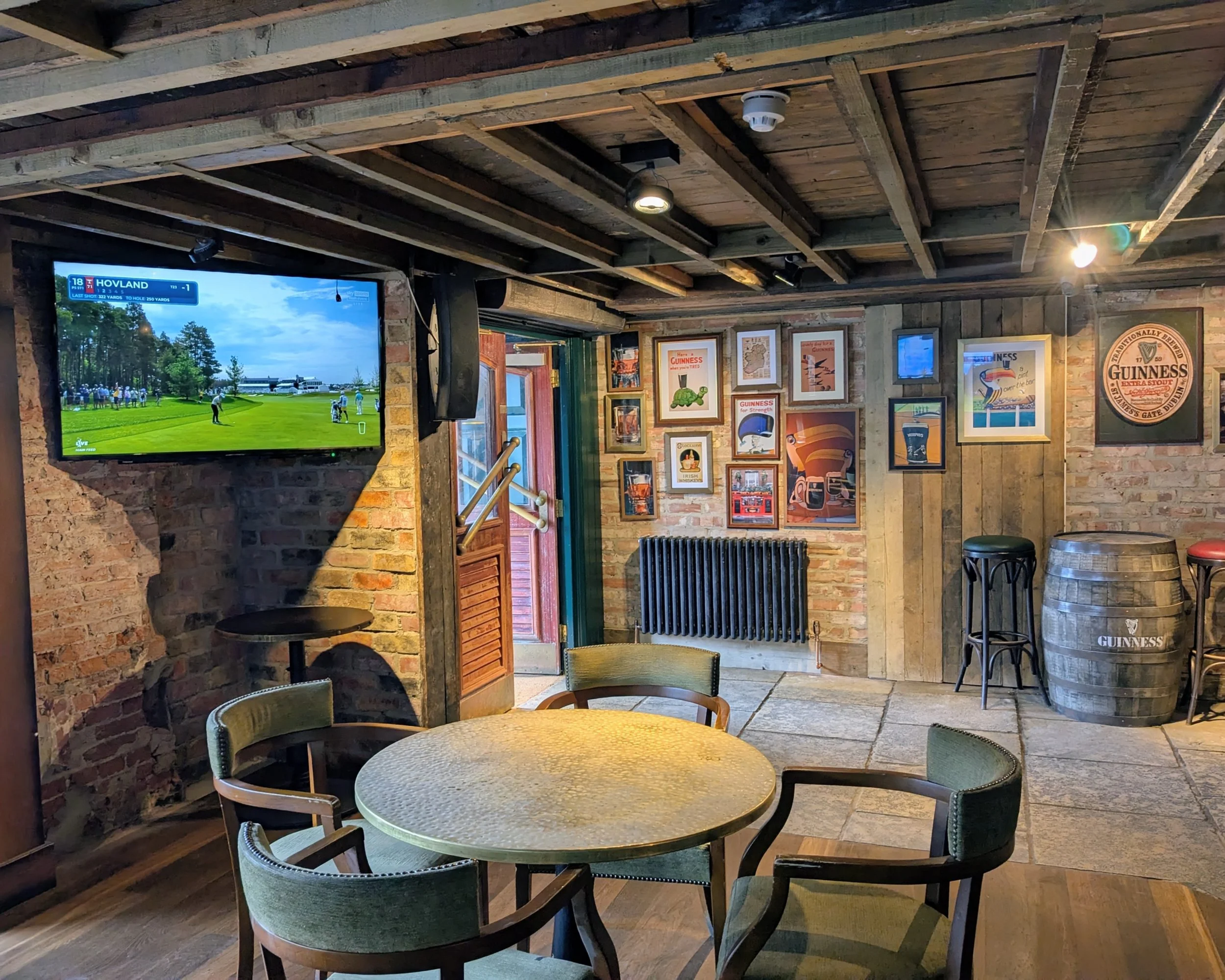 Interior of a rustic pub or restaurant with brick walls and wooden ceiling beams, featuring framed pictures and posters, a flat-screen TV displaying a golf tournament, and a wooden barrel. There are tables and chairs in the space.