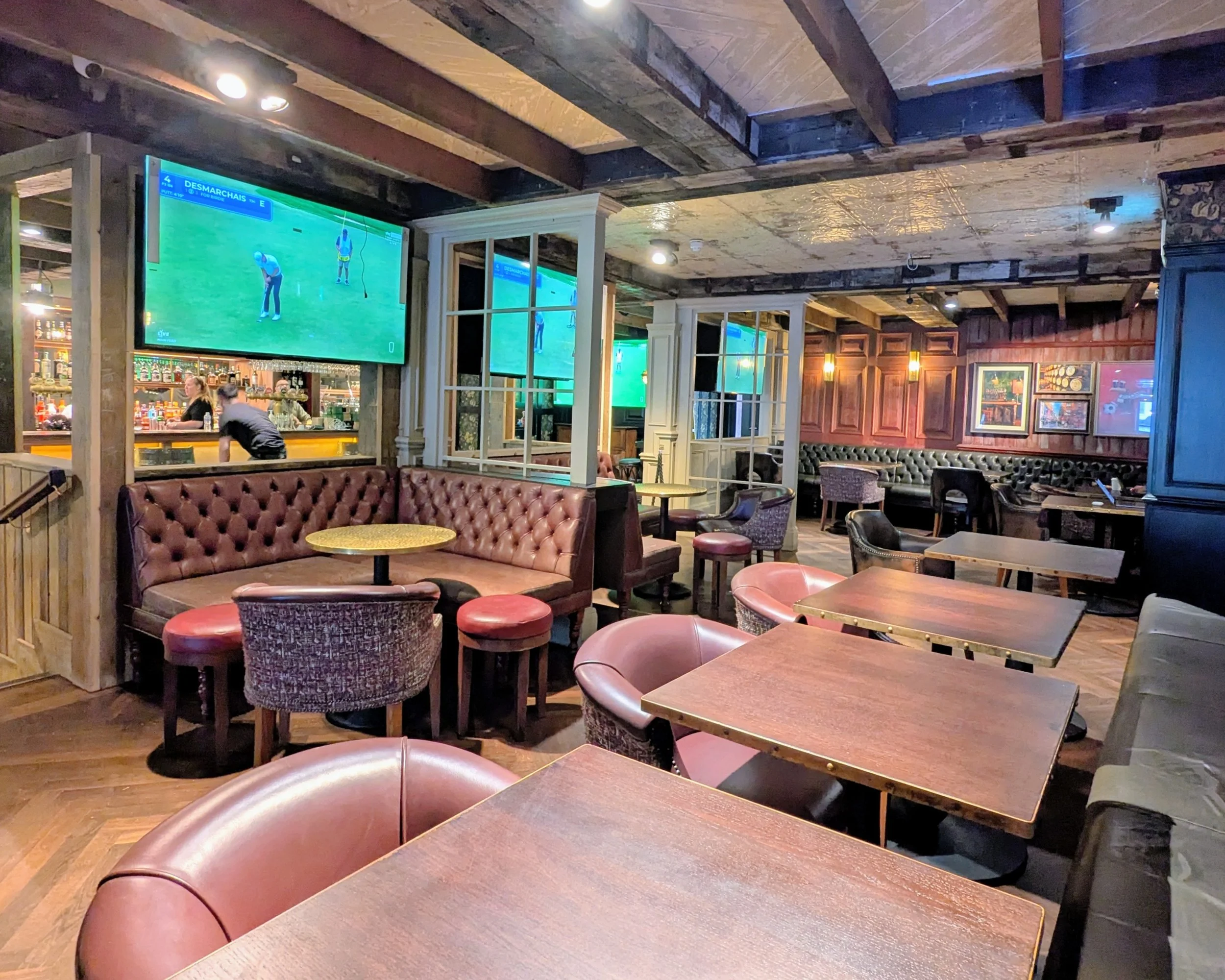 Interior of a bar or restaurant with wooden ceiling beams, multiple television screens showing a golf game, a countertop bar area, and empty tables and chairs.
