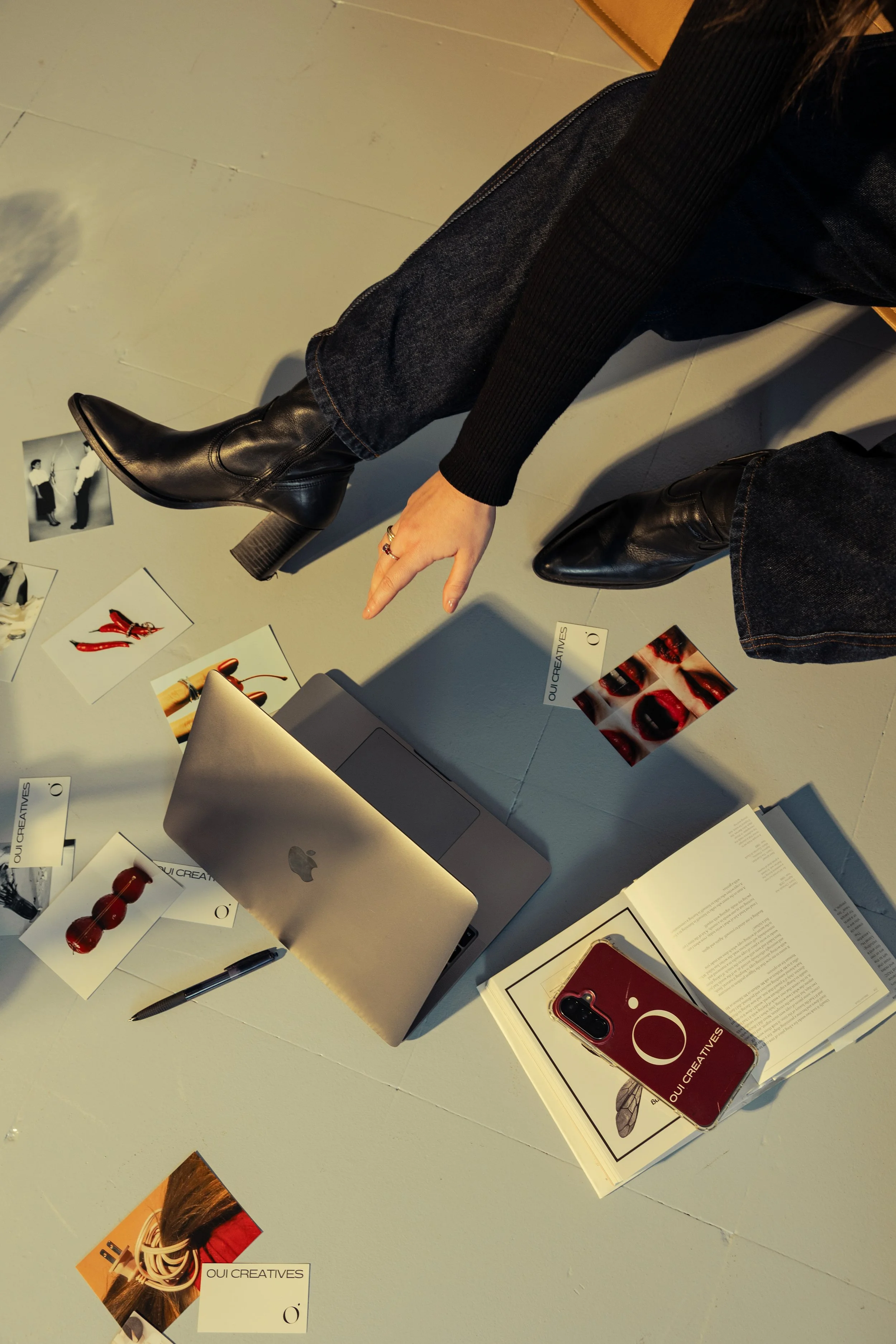 A person wearing black leather boots, dark jeans, and a black sweater is sitting at a desk with various items including a laptop, a smartphone with a red case, printed photographs, printed documents, a pen, and promotional cards for 'OUI CREATIVES'. The environment appears to be an office, workspace, or a creative studio.