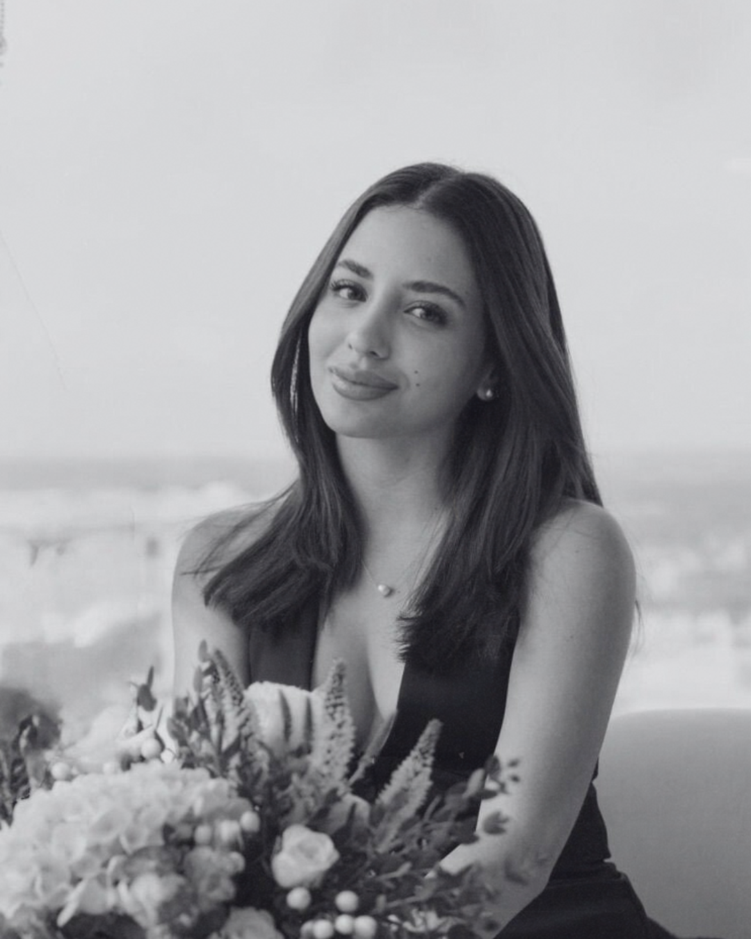 A woman with long dark hair and a necklace, sitting with a bouquet of flowers in front of her, outdoors on a bright day.