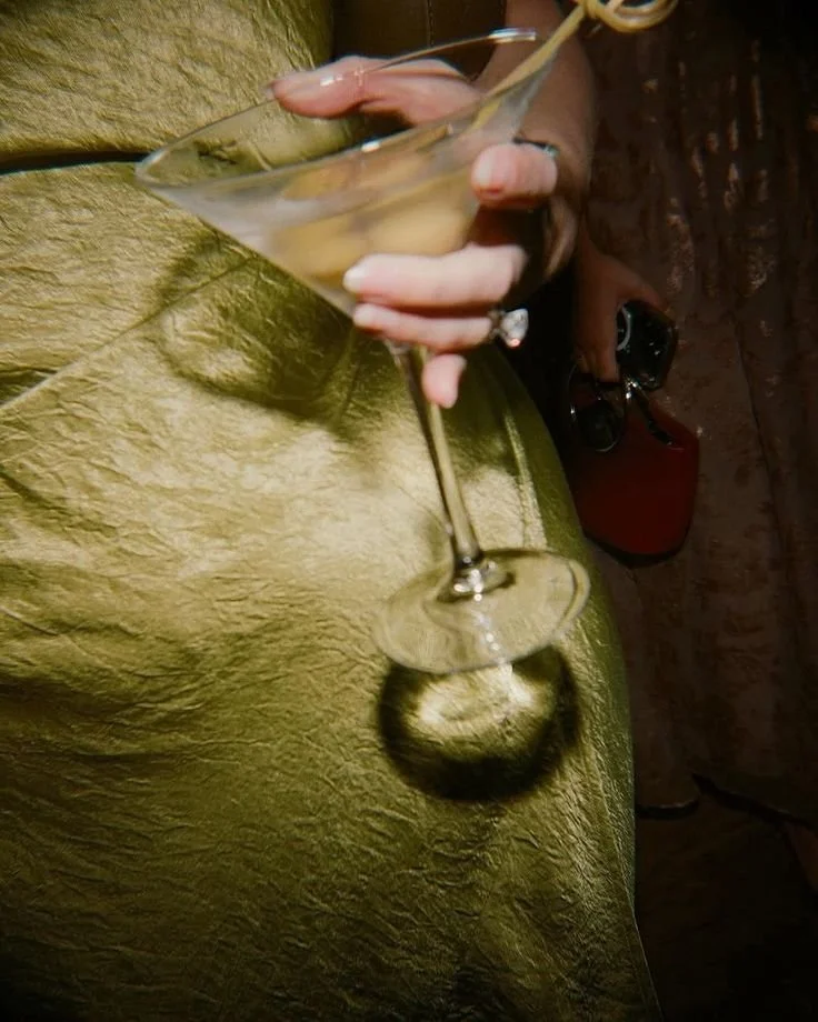 Person holding a cocktail glass with a light-colored drink, sitting on a textured green surface, with sunglasses and a red object in the background.