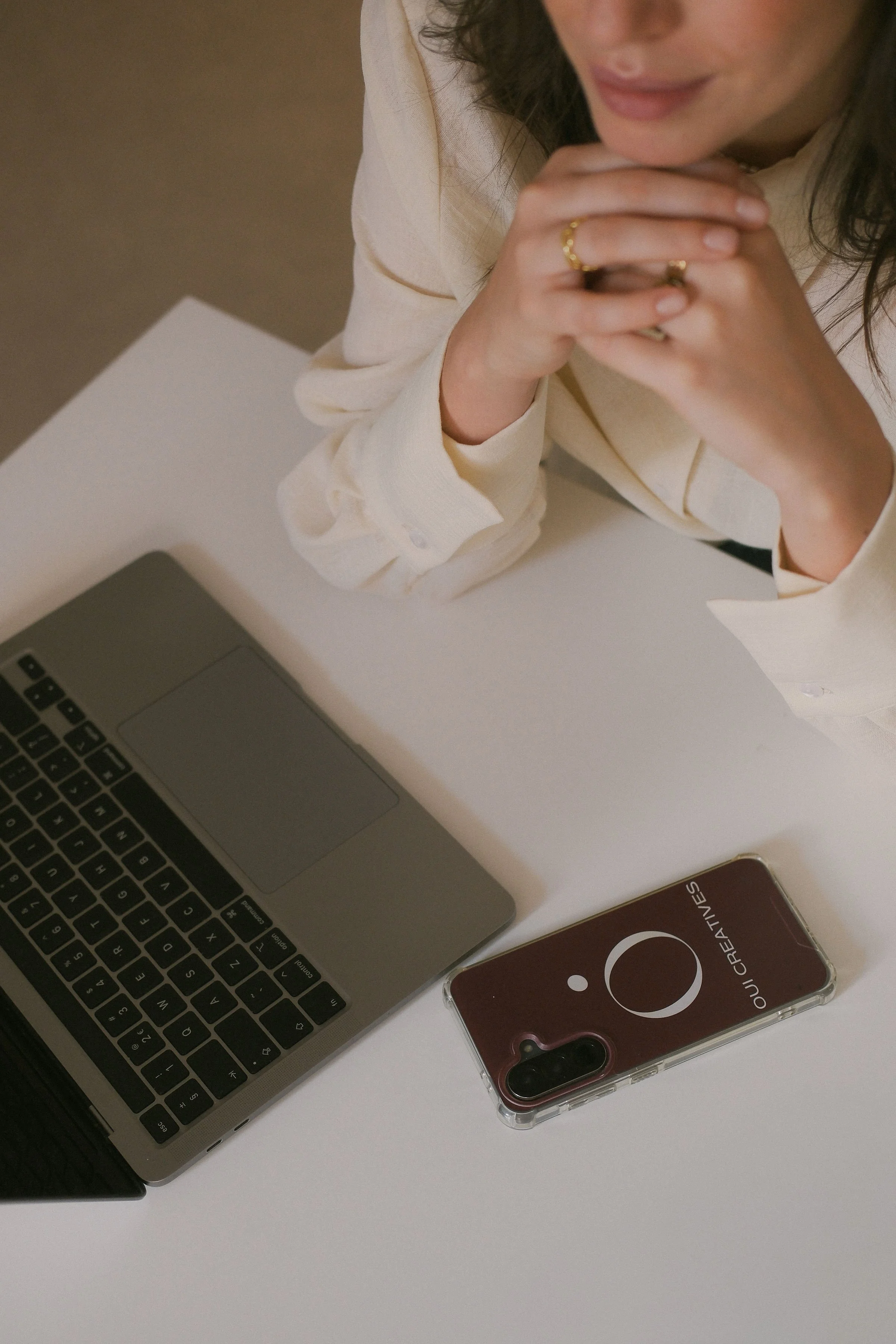 A woman sitting at a white desk with a MacBook laptop and a brown iPhone case on it, wearing a light-colored blouse and gold jewelry. Lodovica Toffoletto FOUNDER OF OUI CREATIVES