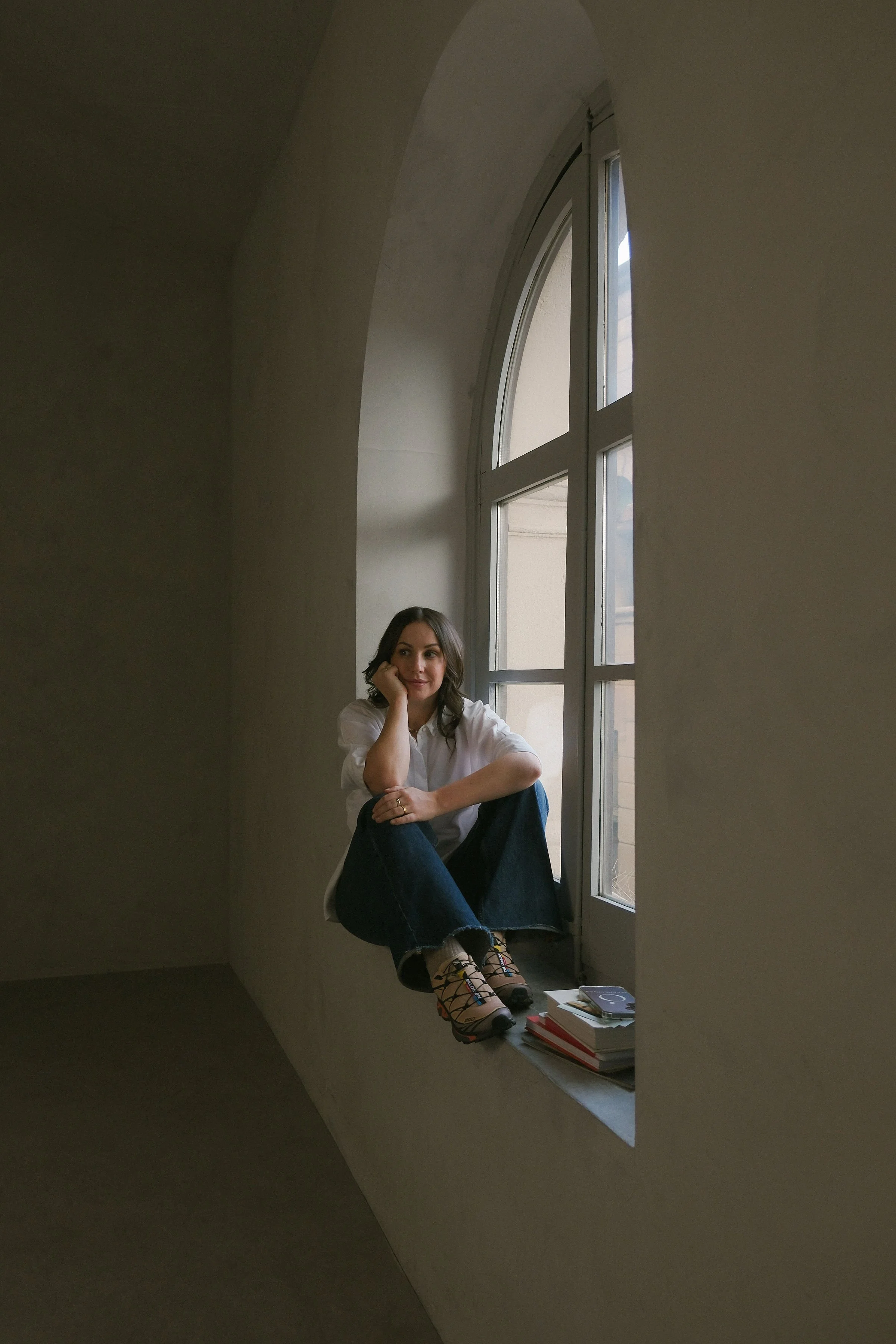 A woman sitting on a window ledge in a room with a large arched window, reading material stacked beside her, wearing a white shirt, jeans, and colorful shoes. Lodovica Toffoletto FOUNDER OF OUI CREATIVES