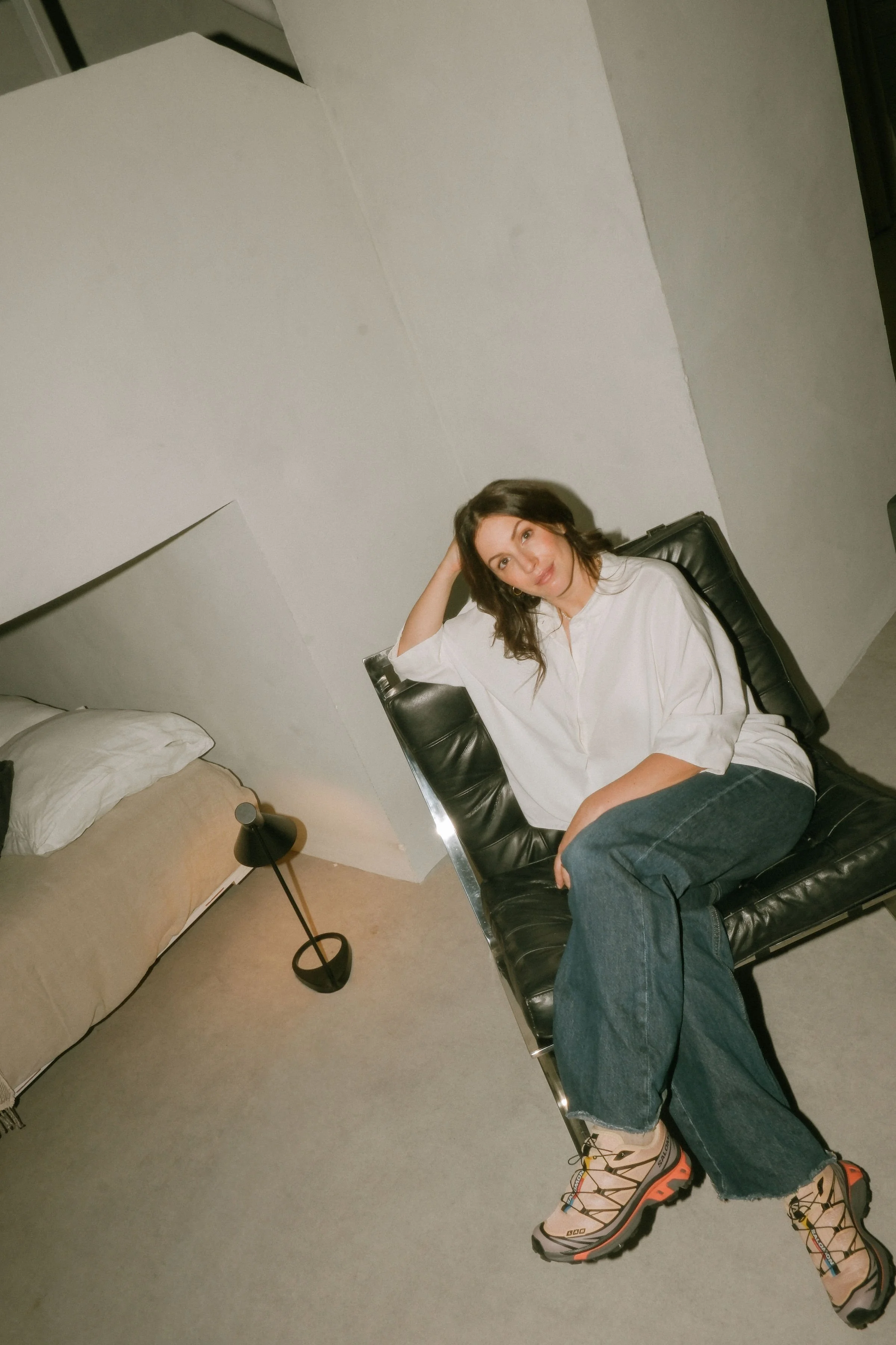 A woman with shoulder-length brown hair in a white oversized shirt, dark wide-leg jeans, and beige hiking shoes sitting on a black leather chair in a minimalist room Lodovica Toffoletto FOUNDER OF OUI CREATIVES