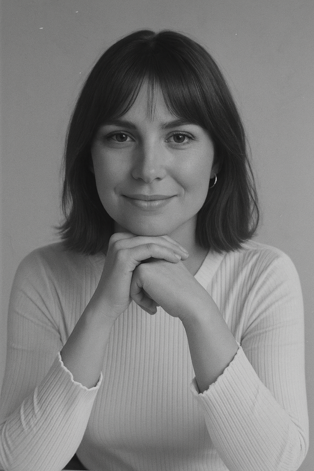 Black and white portrait of a woman with shoulder-length hair and bangs, smiling, with her hands gently clasped under her chin, wearing a long-sleeve ribbed top.