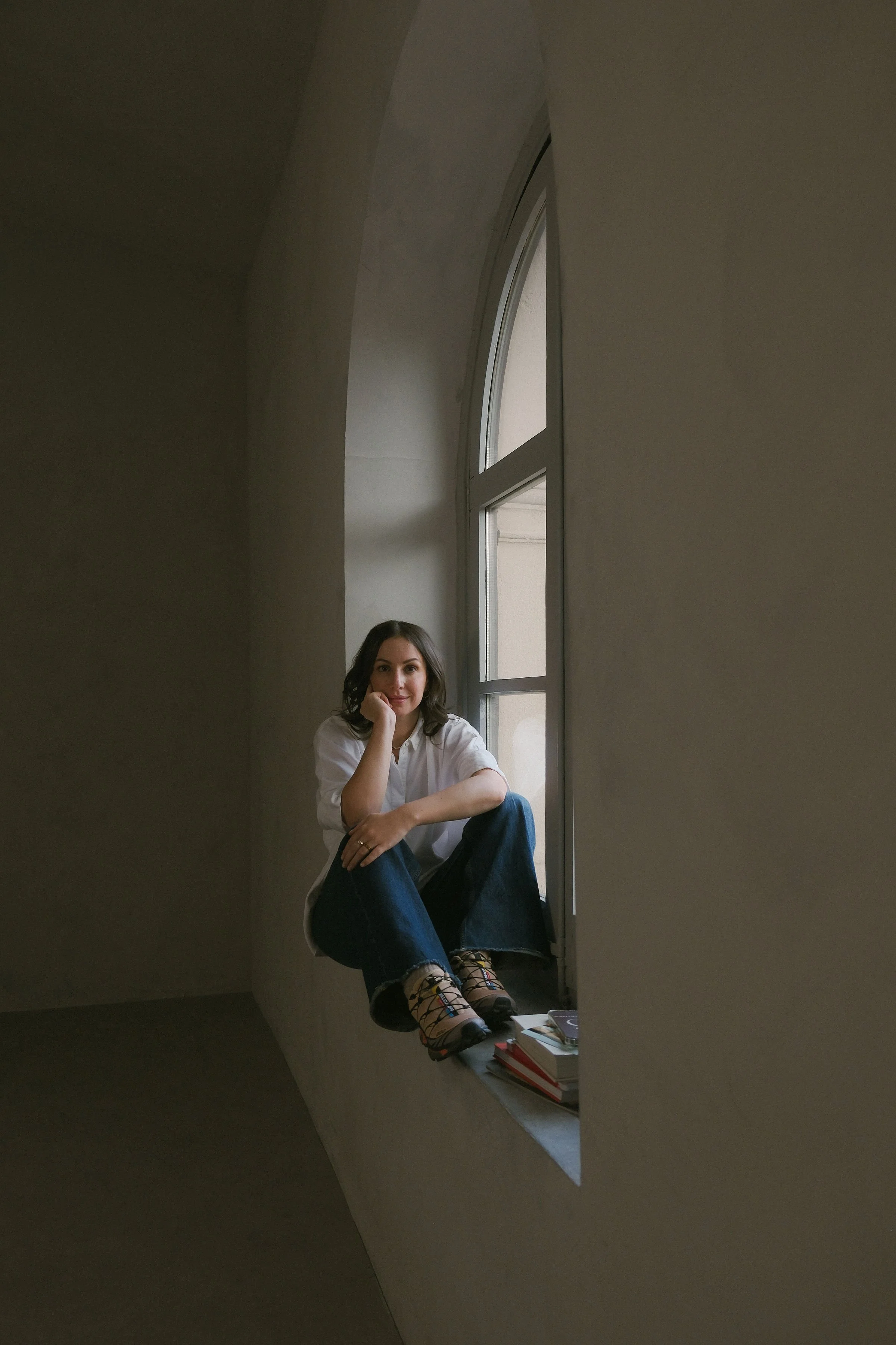 A woman sitting on a wide window ledge, looking at the camera, with her chin resting on her hand, near a large arched window. Lodovica Toffoletto FOUNDER OF OUI CREATIVES