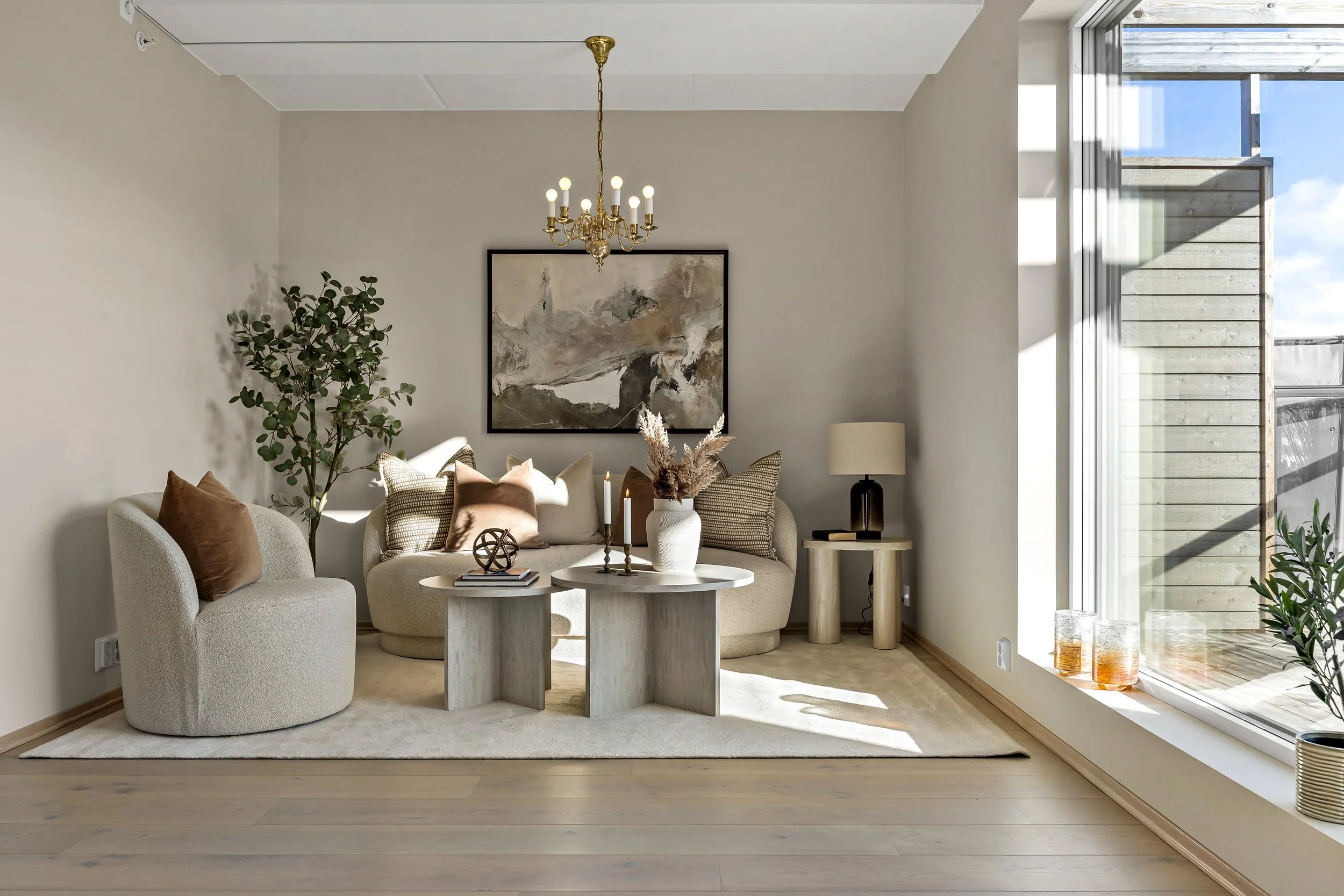 Stylet stue med beige sofa og stol, to hvite bord, blomsterpotter, kunst på veggen og store vinduer med utsikt.