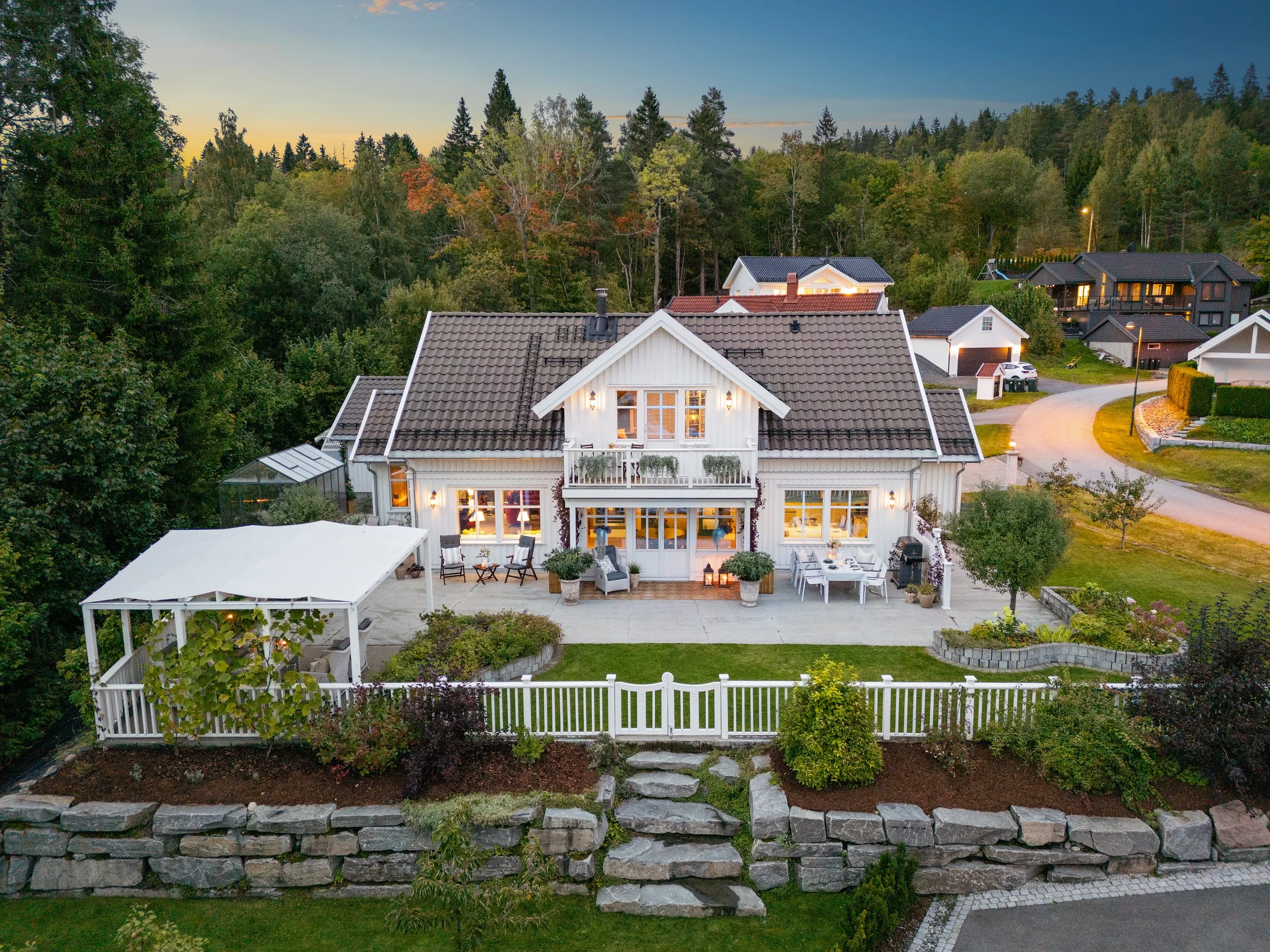 Et koselig kveldsbilde av et hvitt hus med opplyste vinduer og terrasse, omgitt av grøntområder og trær om kvelden. Varme farger i horisonten.