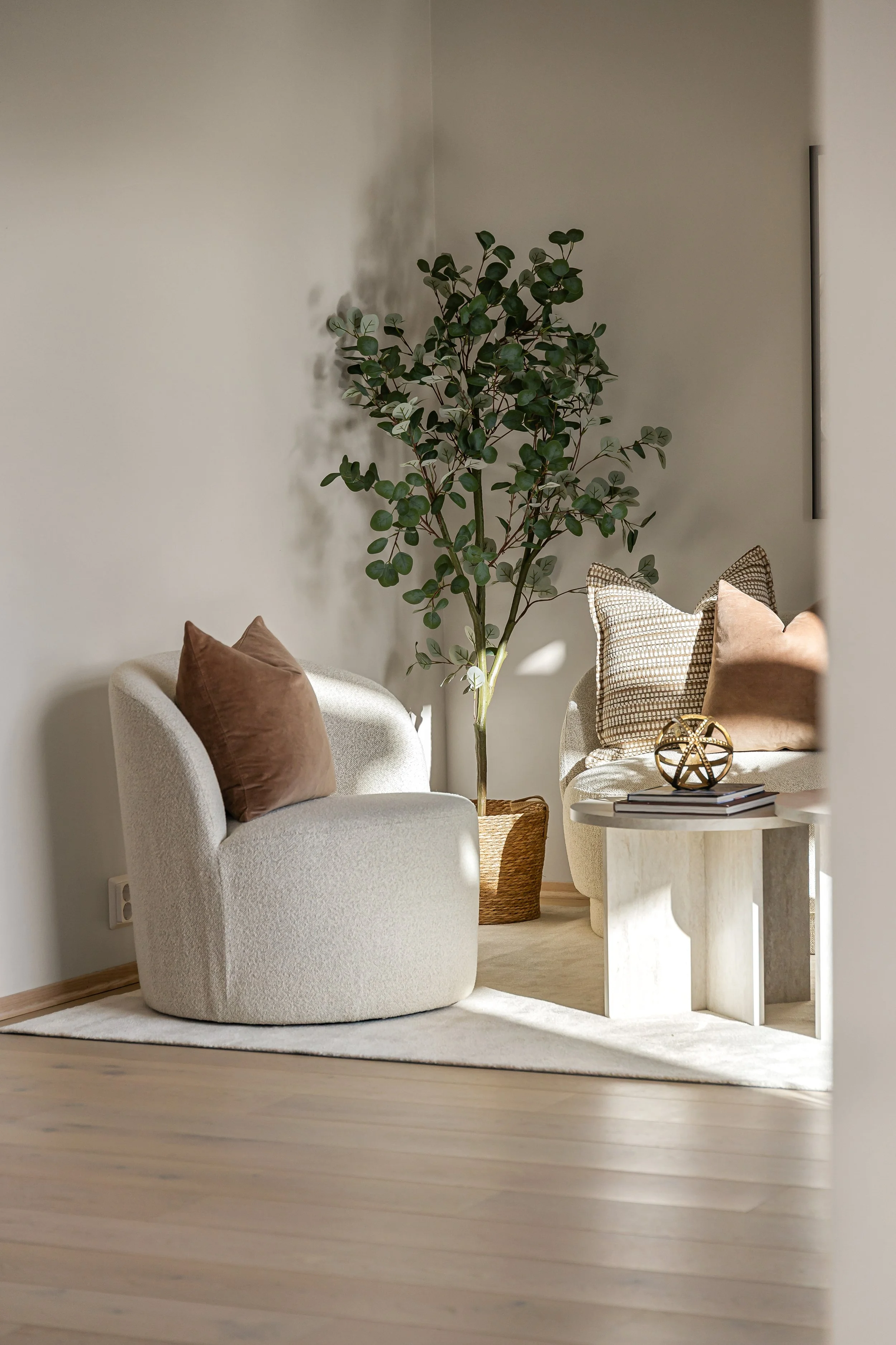 Lys stue med en hvit, rund sofa med brune puter, en stor plante i kurv, en hvit sidebord med bøker og dekorative gjenstander, og en liten hvit teppe på tregulvet. Stylet bolig.