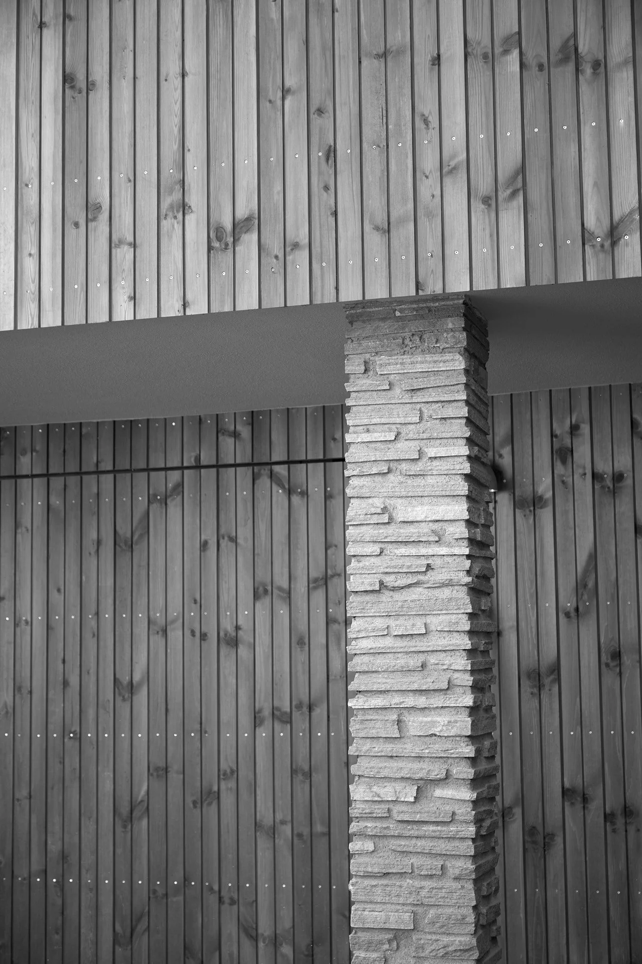 Arkitektonisk utsnitt av bygning med vegger av vertikalt montert trepanel og en stolpe av kløvet stein. Svart-hvitt.