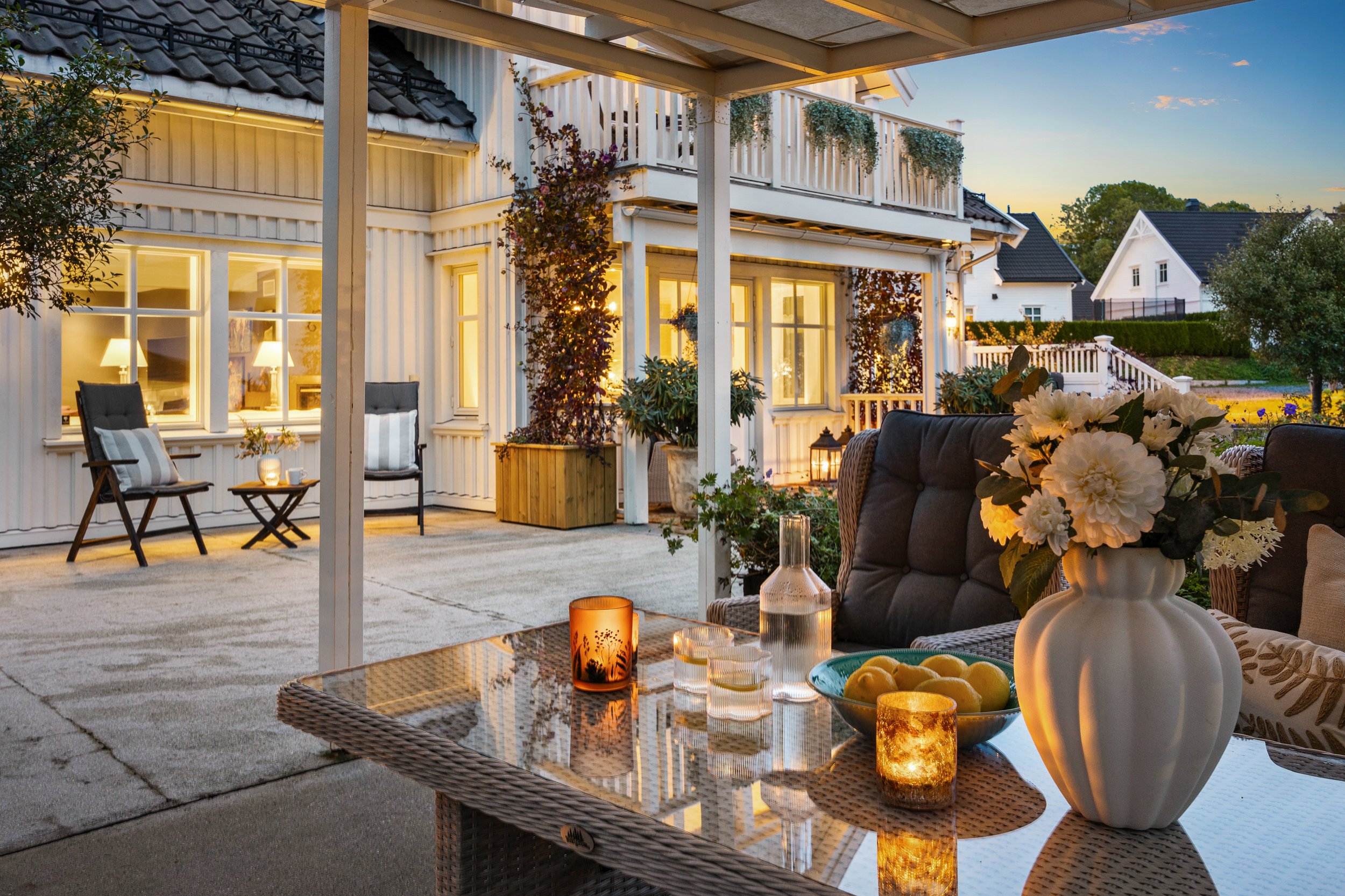 Utendørs terrasse med bord, blomsterbukett i vase, lyslykter og stearinlys, solnedgang i bakgrunnen, uterom med komfortable sitteplasser og planter. Kveldsbilde med varm belysning fra levende lys og interiørbelysning.
