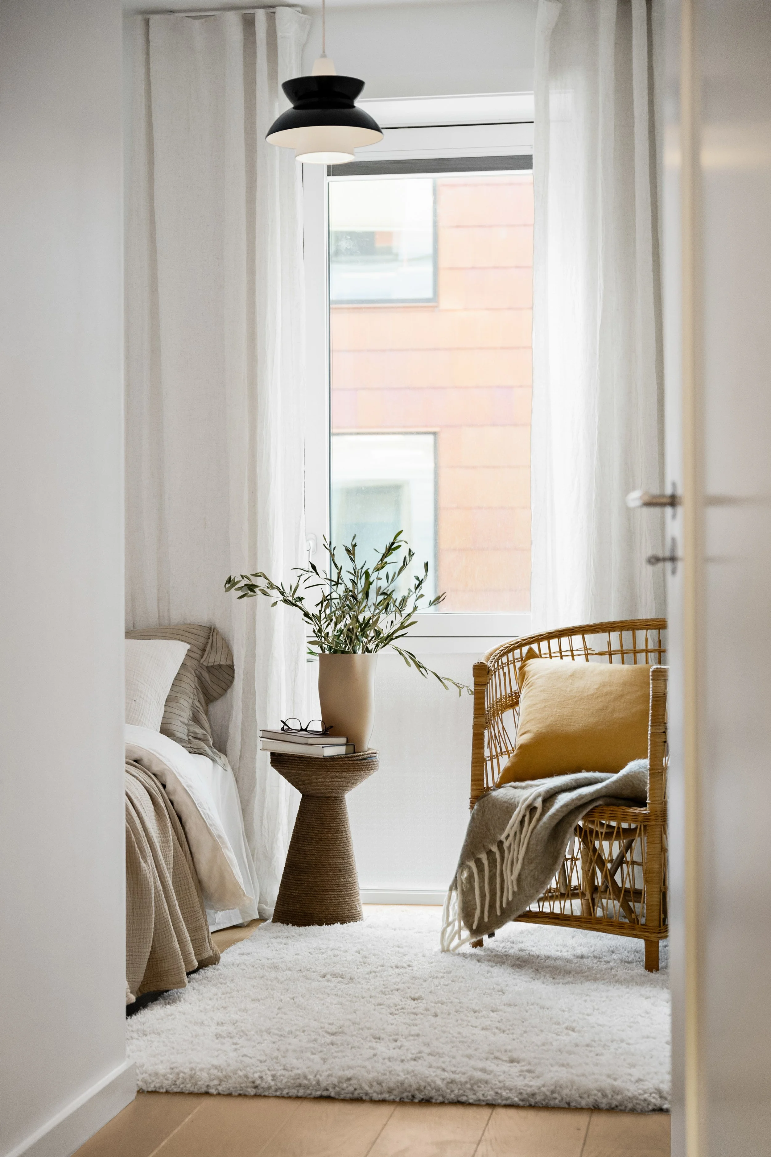 Innbydelig soverom med stort vindu, beige seng med puter, en kurvstol med pute og teppe, et lite sidebord med planter og bøker. Moderne styling.