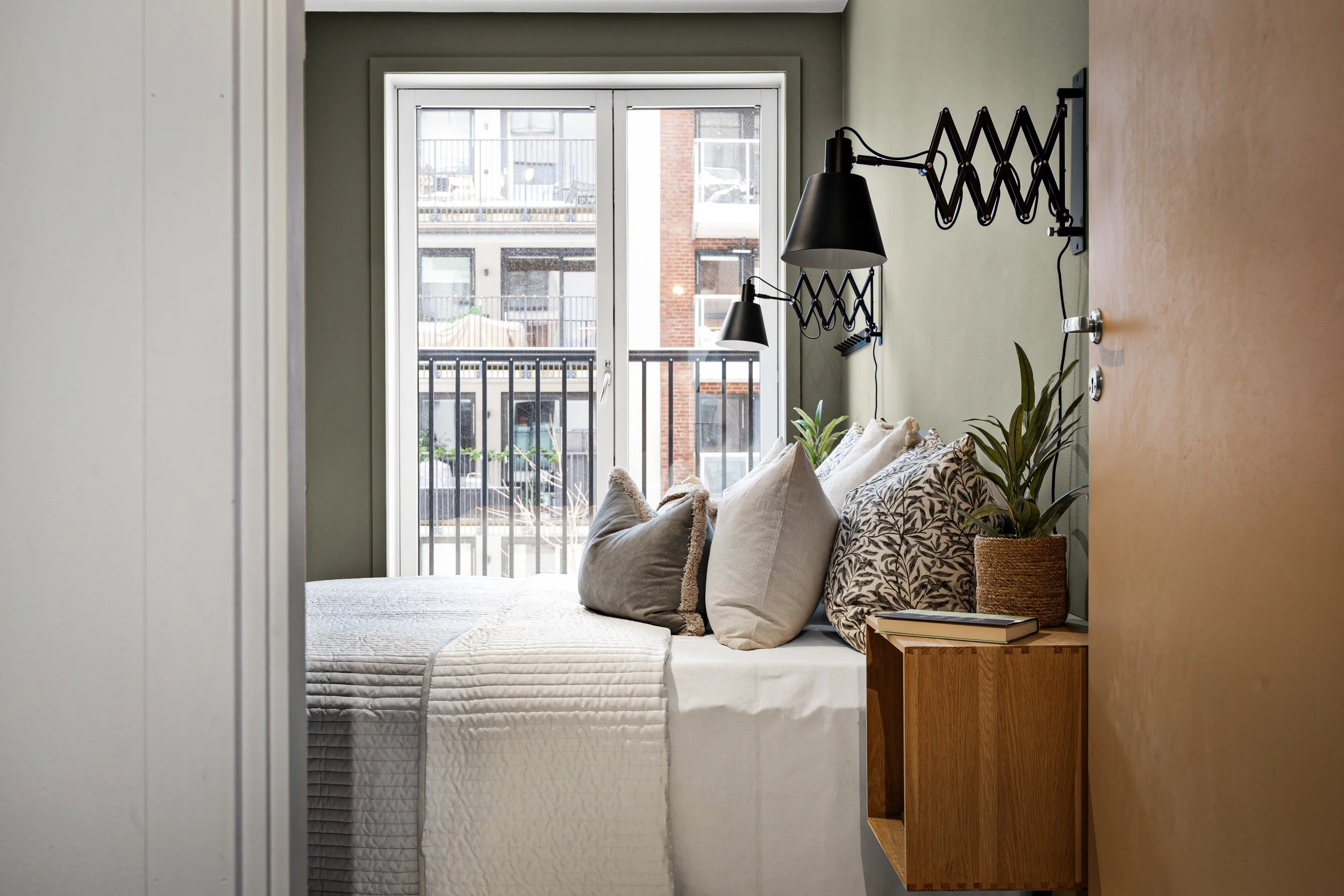 Stylet soveom med seng, puter, vindu, balkong, delikate farger, naturlig lys, vegglamper og plante i keramikkpotte.