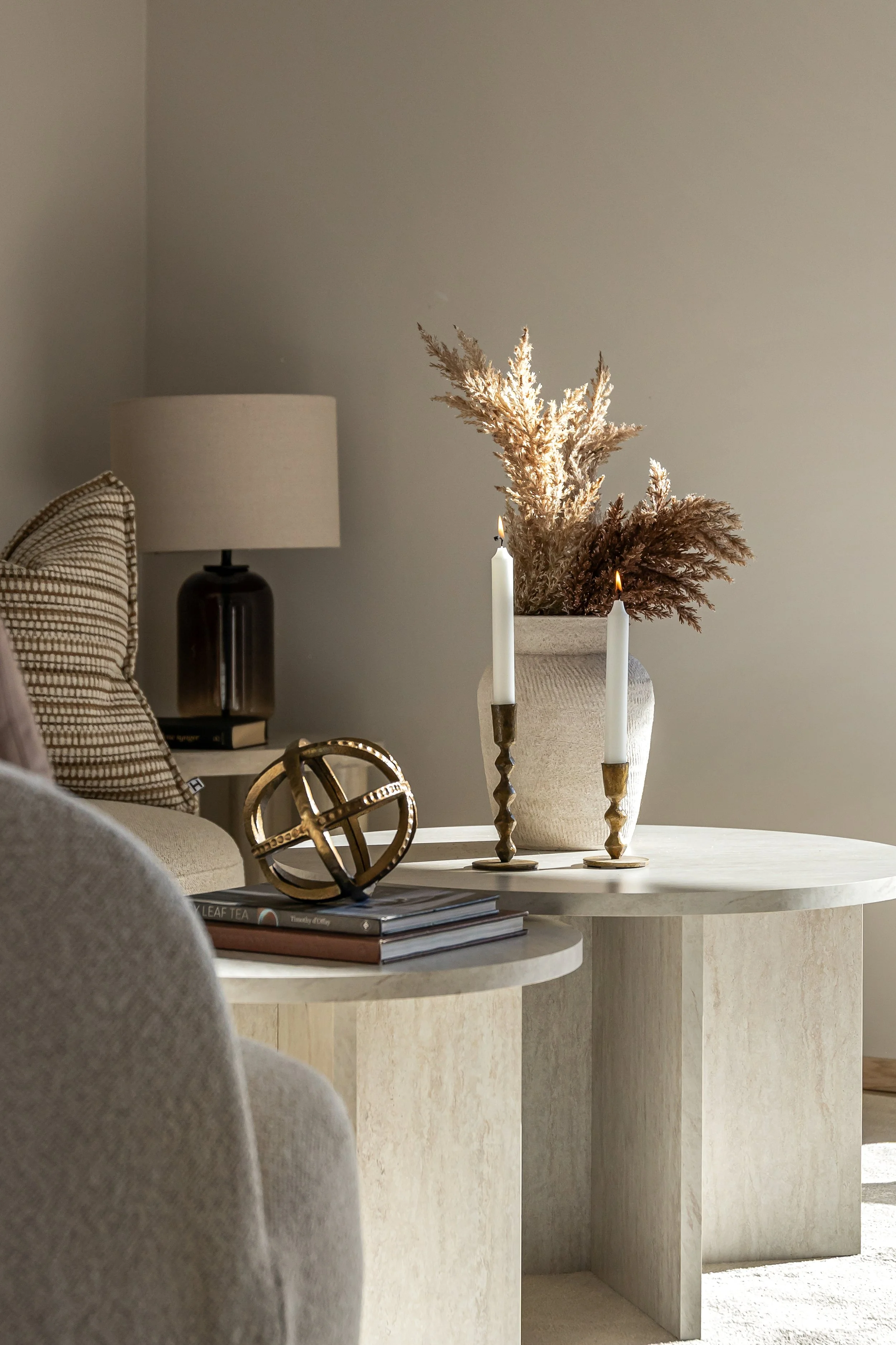 Et rundt spisebord laget av lyst tre med en vase fylt med tørkede blomster, to hvite lysestaker og noen bøker på toppen. En del av en beige sofa og et bordlampe er synlig i bakgrunnen.