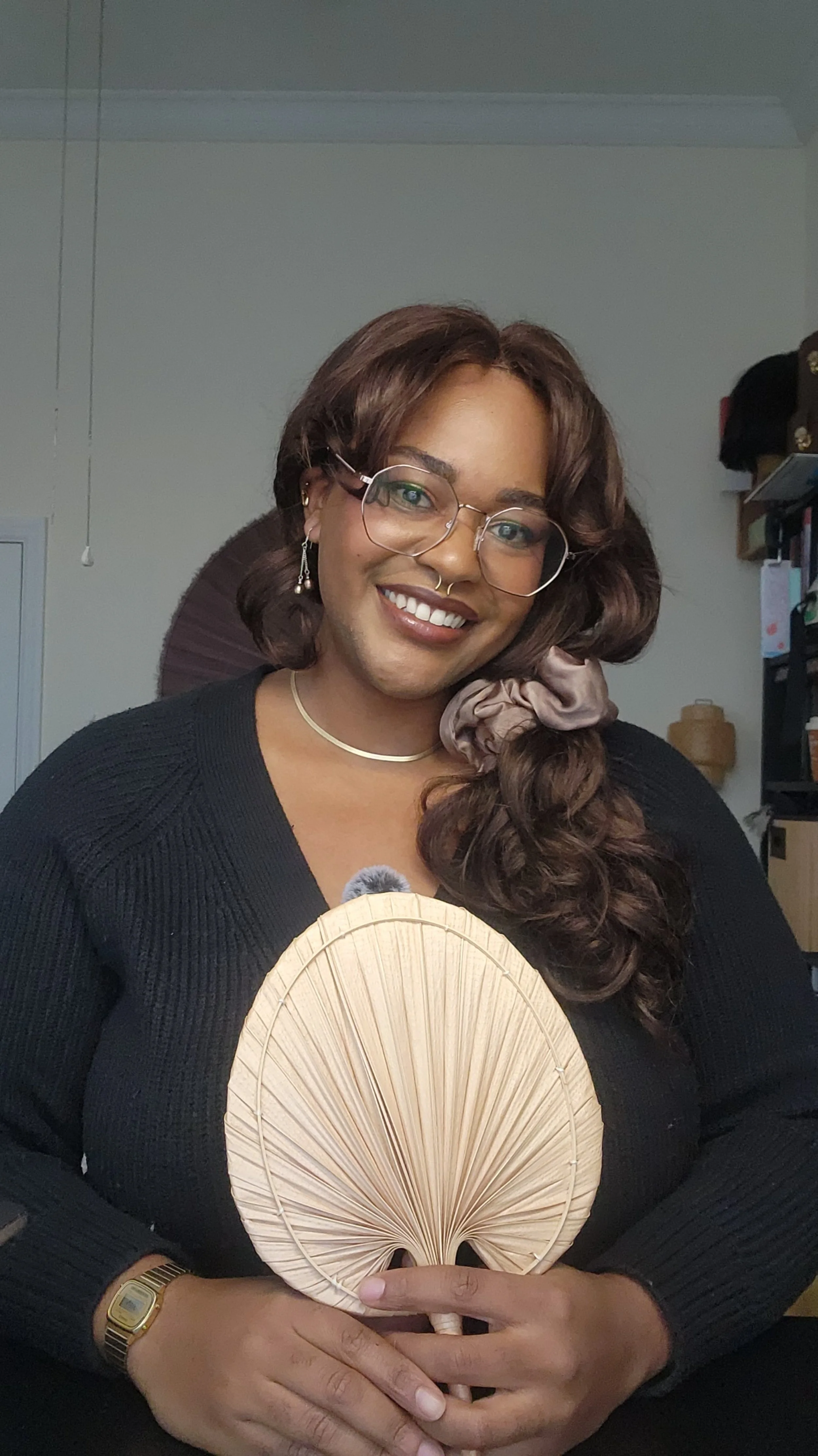 A woman with glasses, brown hair styled in curls with a scrunchie, smiling and holding a round woven fan, dressed in a black top and wearing jewelry, in an indoor setting.