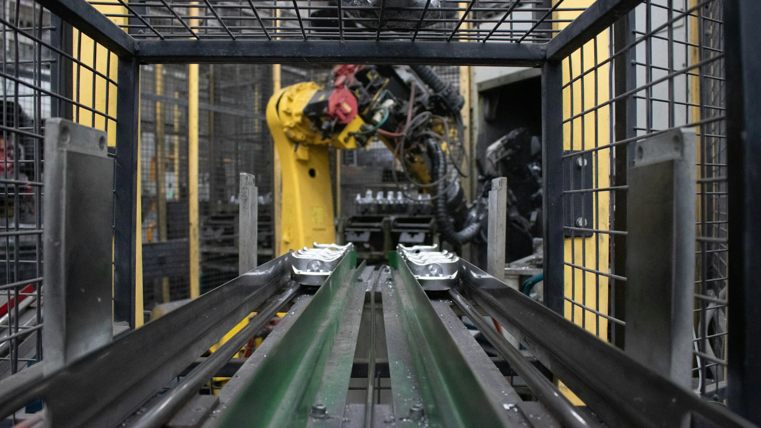 Inside a factory or warehouse with a robotic arm working on conveyor belts, surrounded by metal safety fences.