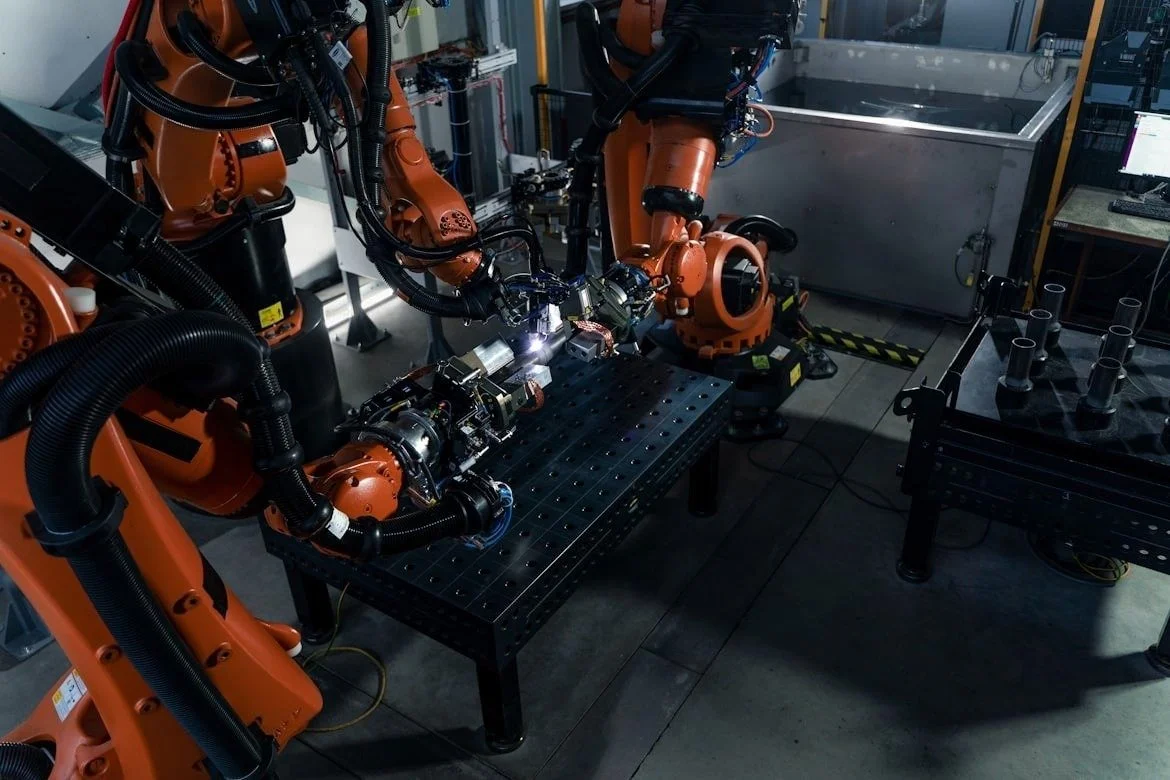 Multiple orange industrial robots working together on a black perforated metal table in a high-tech manufacturing lab, with computer monitors and equipment in the background.