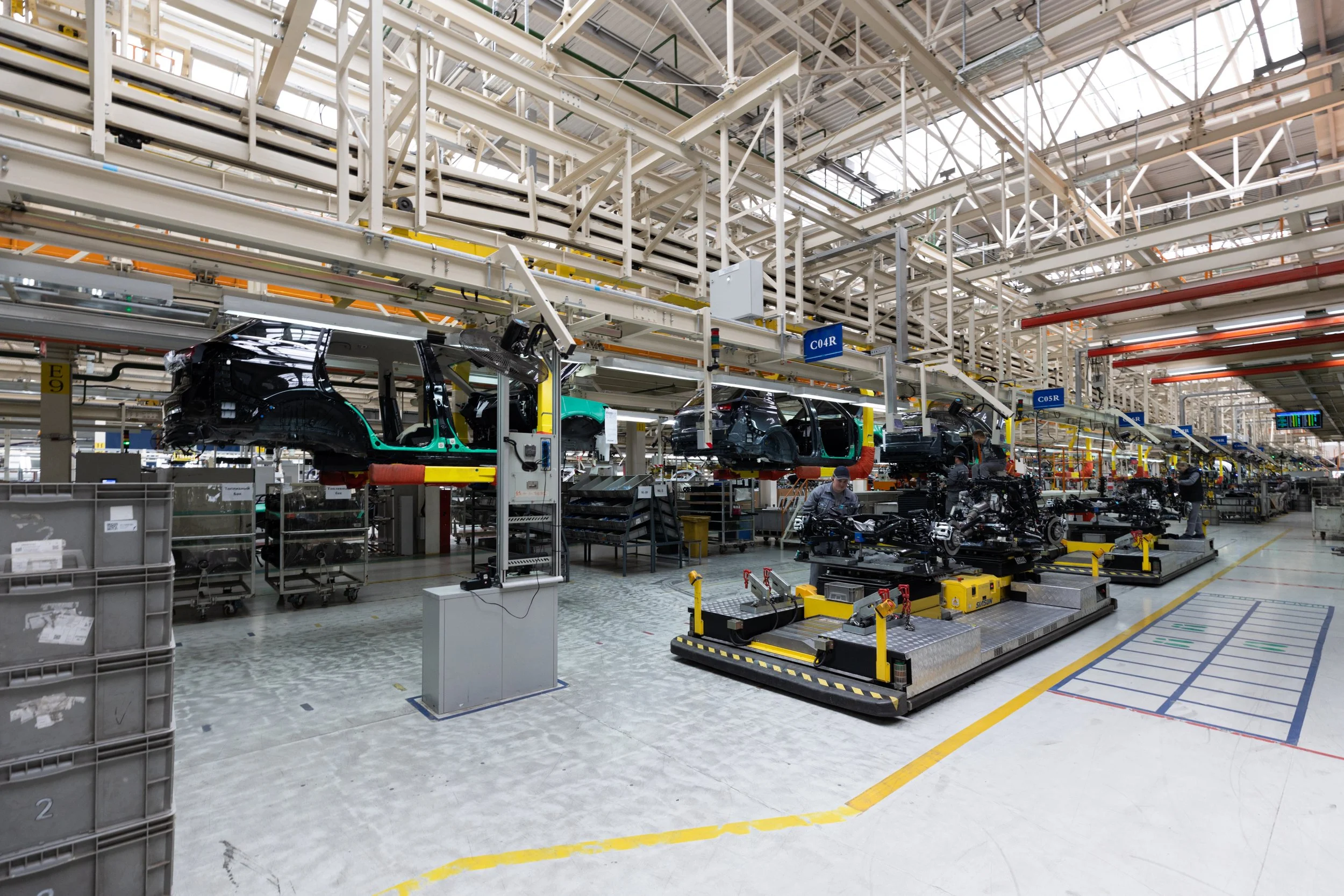 Automobile assembly line inside a factory with car bodies on an overhead conveyor system, workers in the background, and organized tools and parts on the floor.
