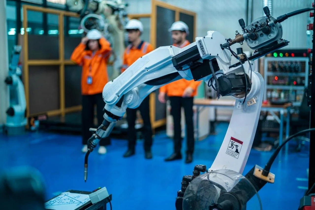 A robotic arm in a laboratory or industrial setting with three workers in safety gear and helmets observing in the background.