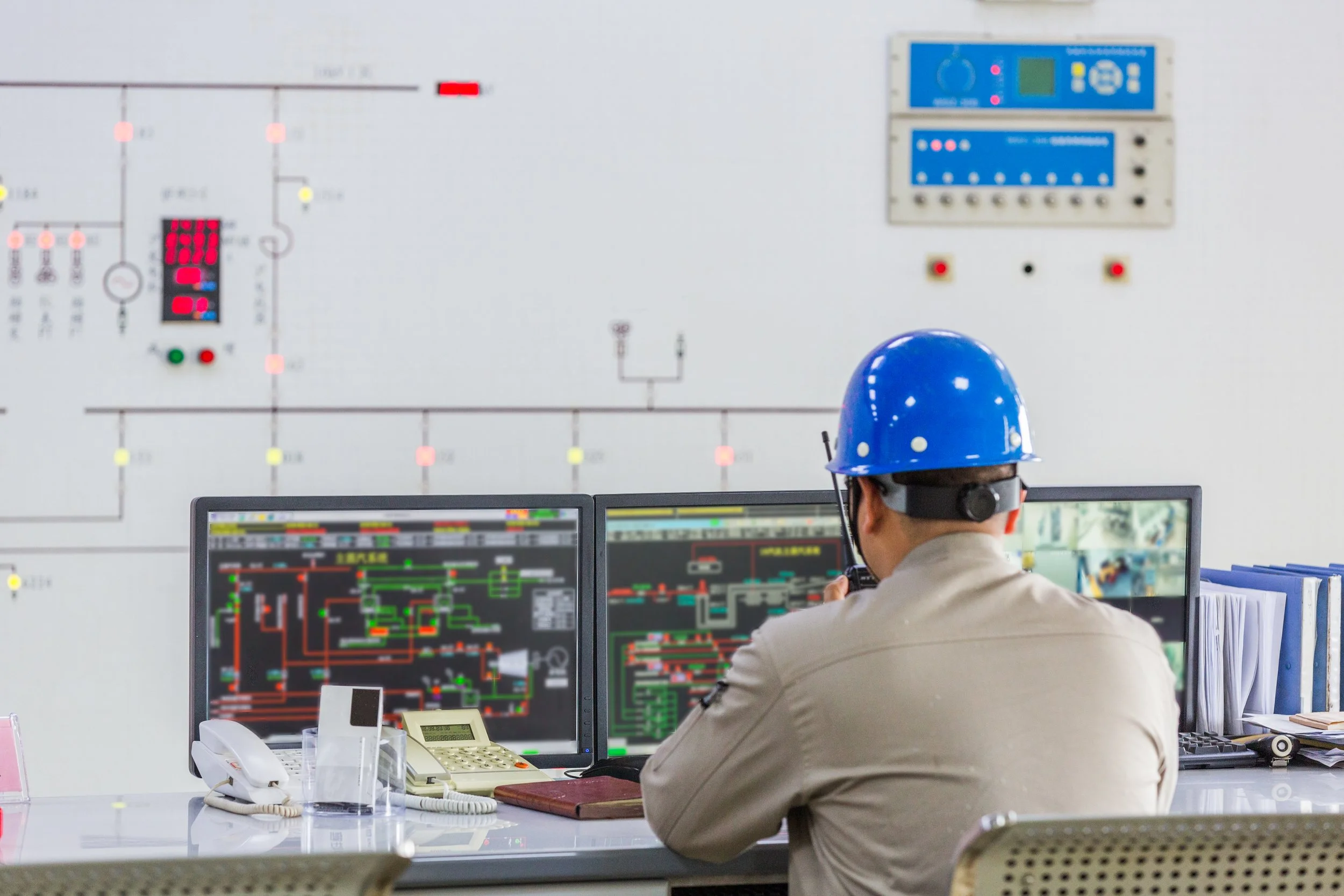 An engineer wearing a blue safety helmet working at a control station with multiple computer screens displaying circuit diagrams and control panels in an industrial setting.