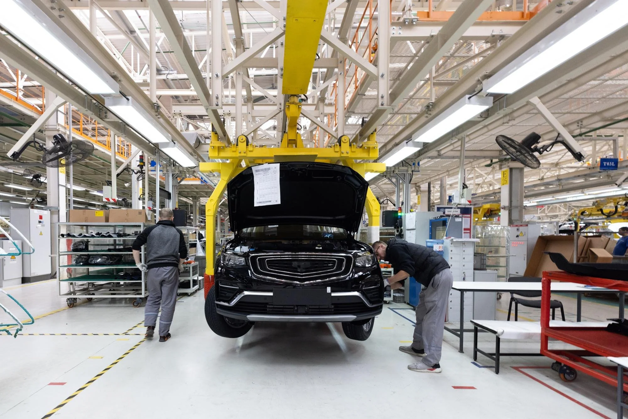 Workers in an automotive manufacturing plant assembling a black SUV with an open hood, surrounded by machinery, tools, and industrial lighting.