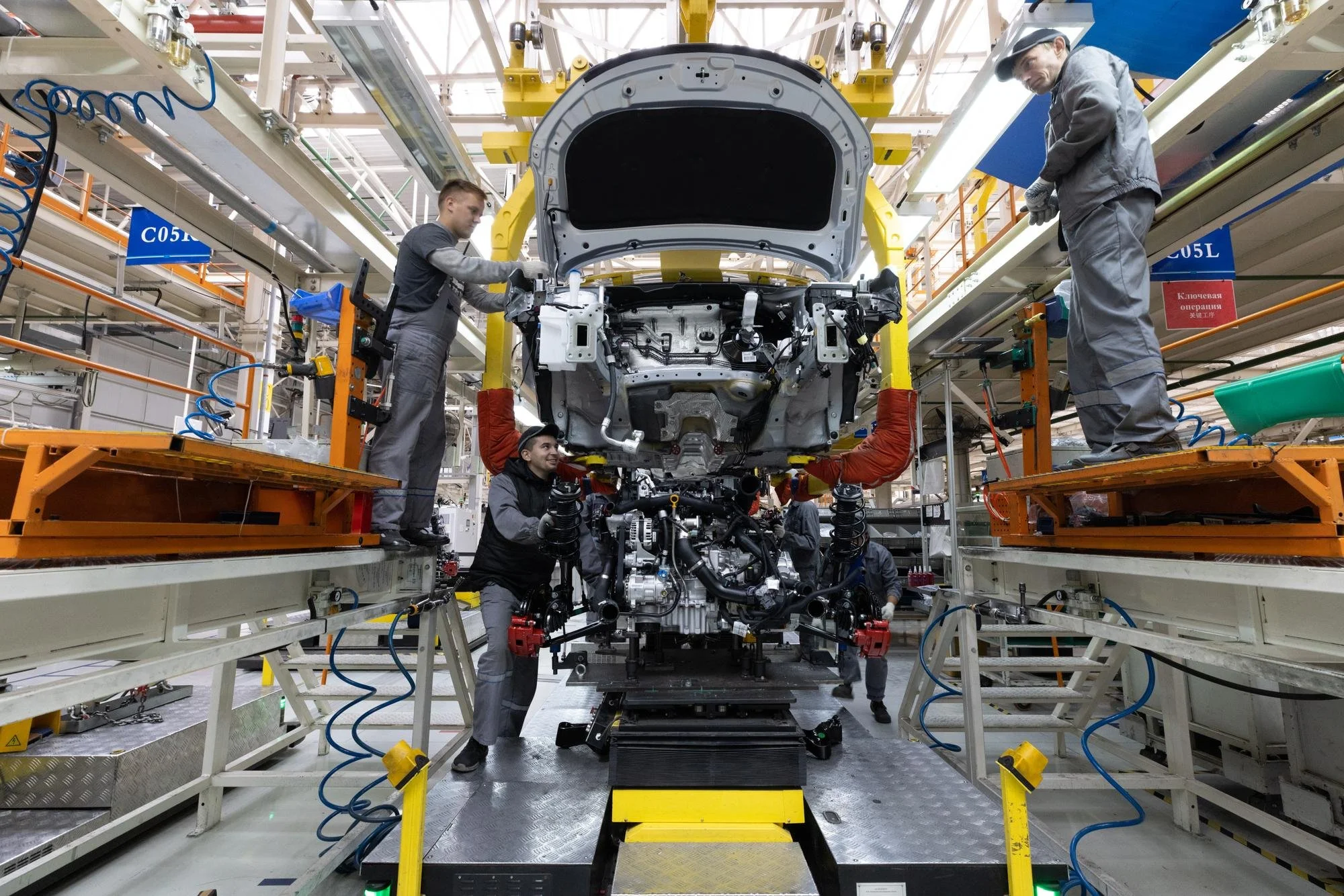 Workers assembling a car body in an automotive manufacturing plant, with robotic and mechanical components surrounding them.