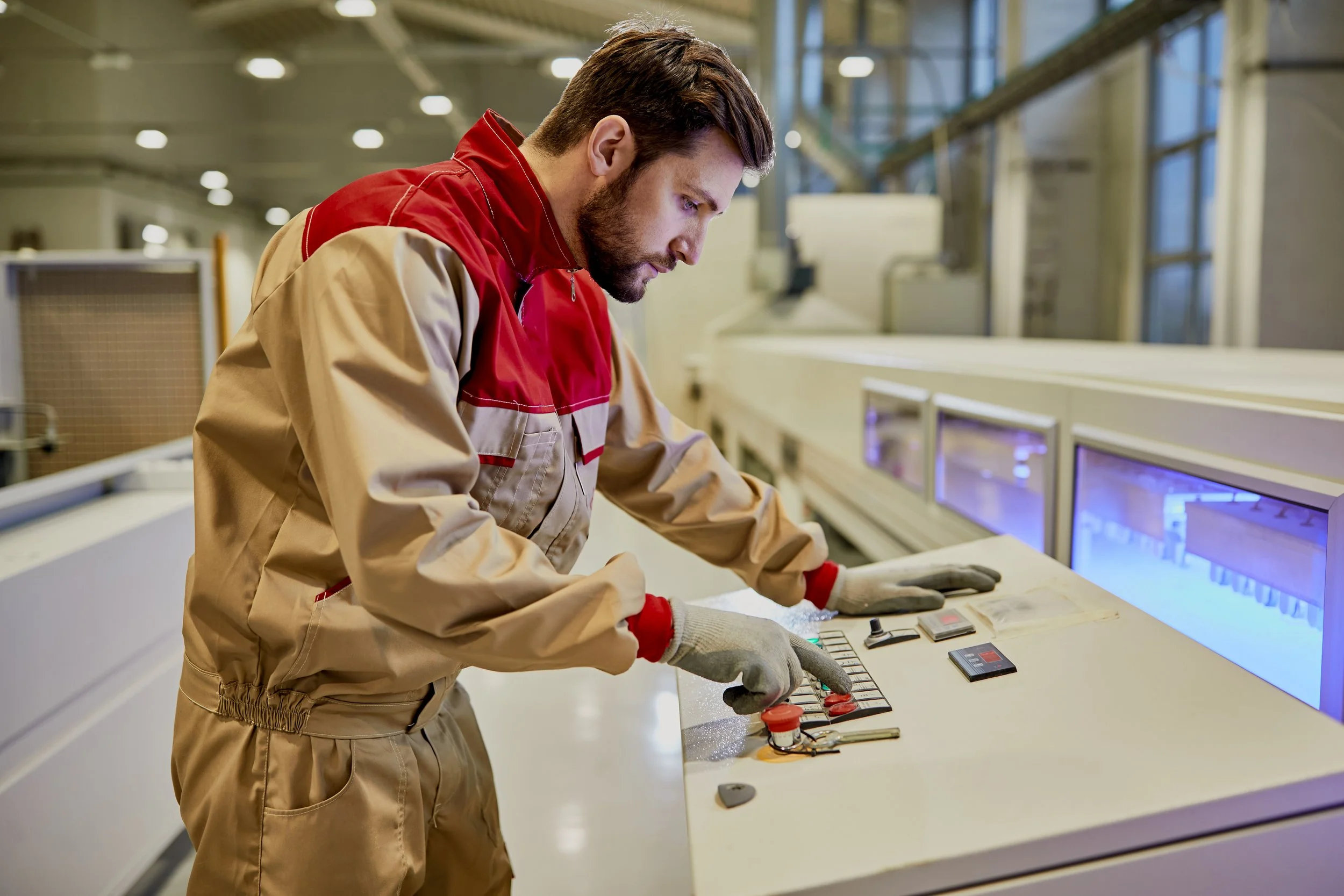 A man wearing a beige and red work uniform is working with electronic components at a workstation in a factory or lab environment, with machinery and windows in the background.