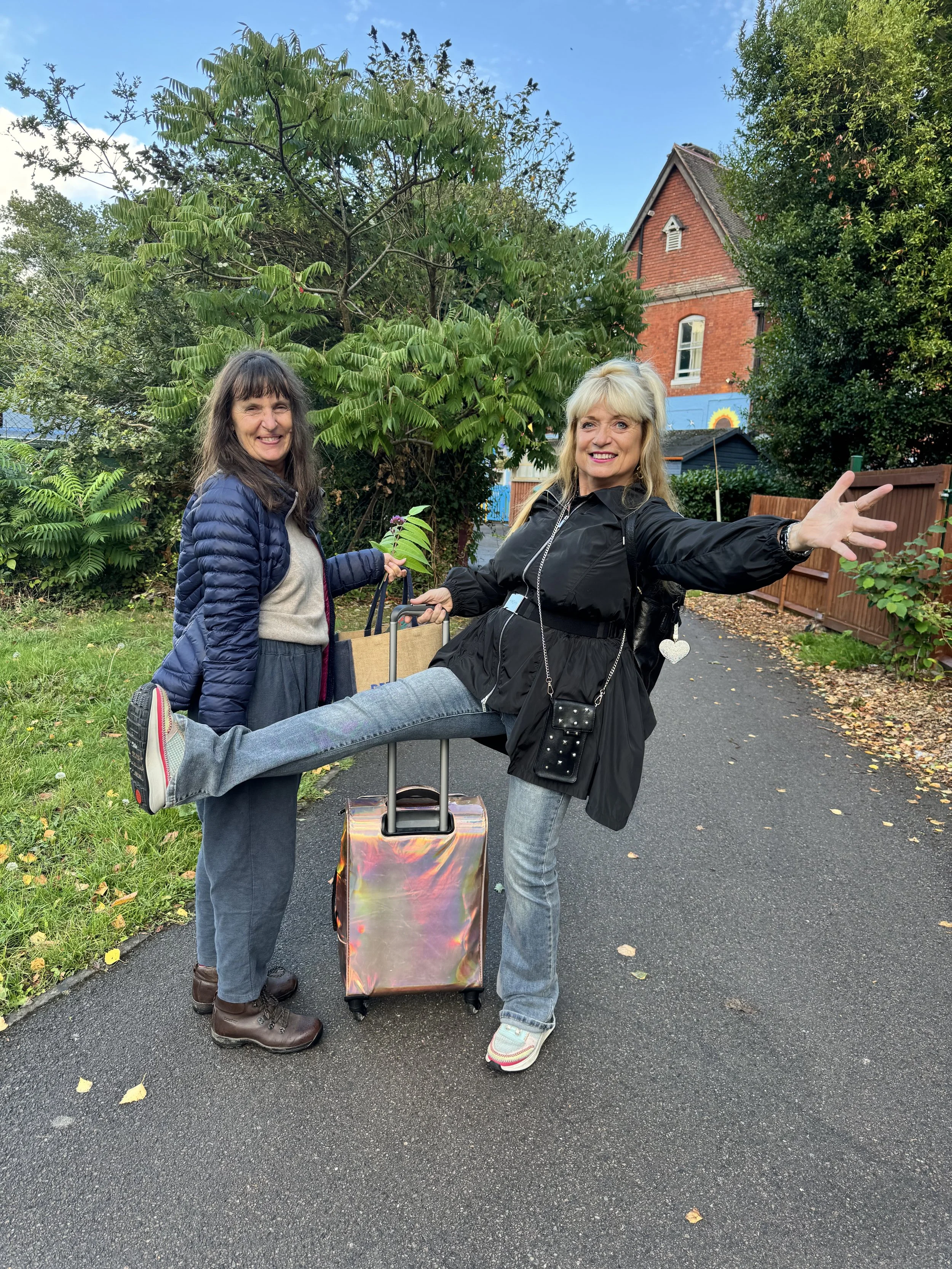 Two women stand on a paved path outdoors, one smiling and holding a plant, the other extending an arm with a playful expression, with a colorful house and green trees in the background.