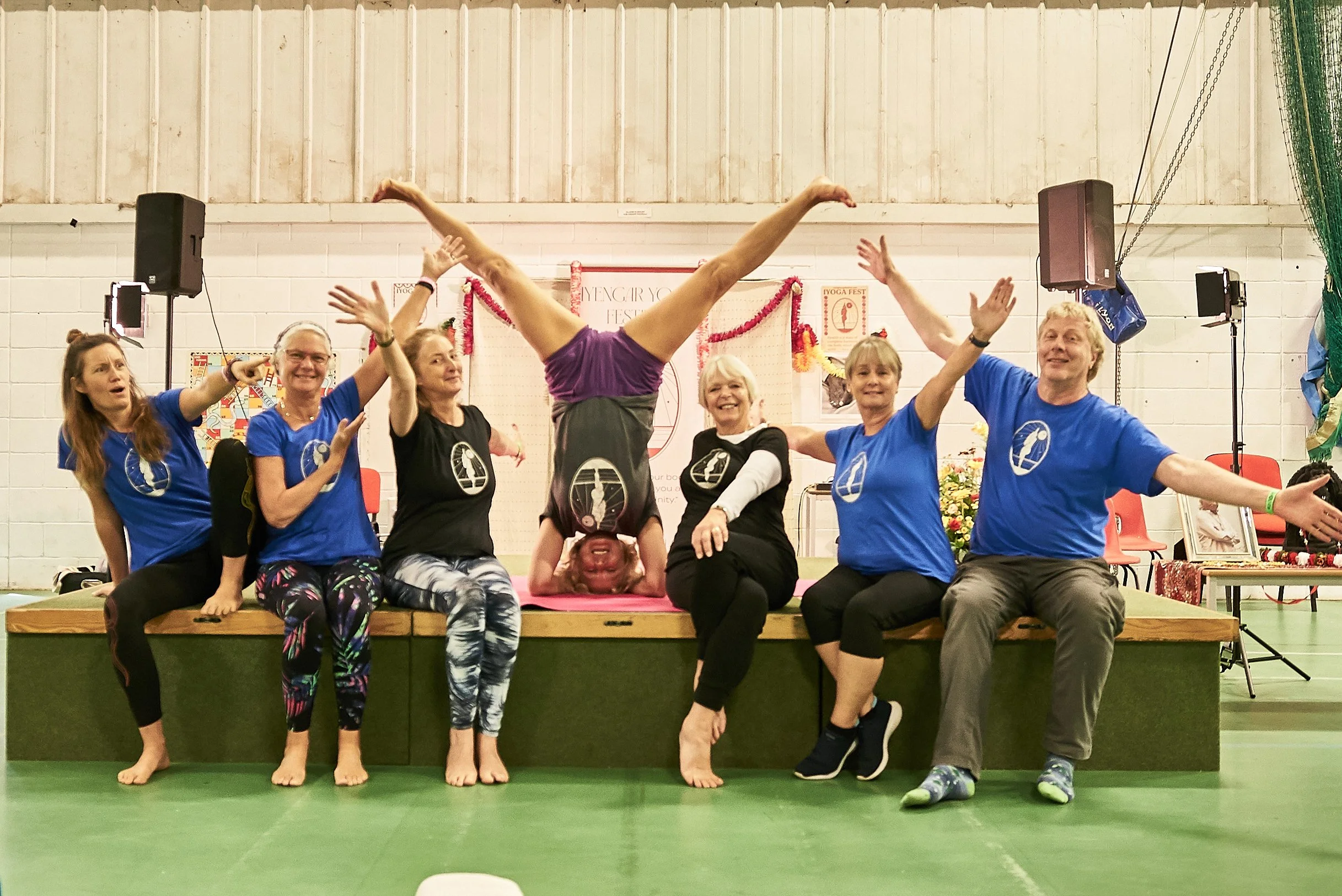 Group of seven people, six women and one man, sitting and one woman doing a headstand, on a stage in a gymnasium or community center, celebrating or performing a yoga or fitness event, with decorations and banners in the background.