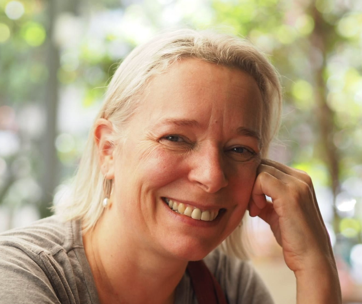 Smiling woman with blonde hair and pearl earrings sitting outdoors with blurred trees and sunlight in the background.
