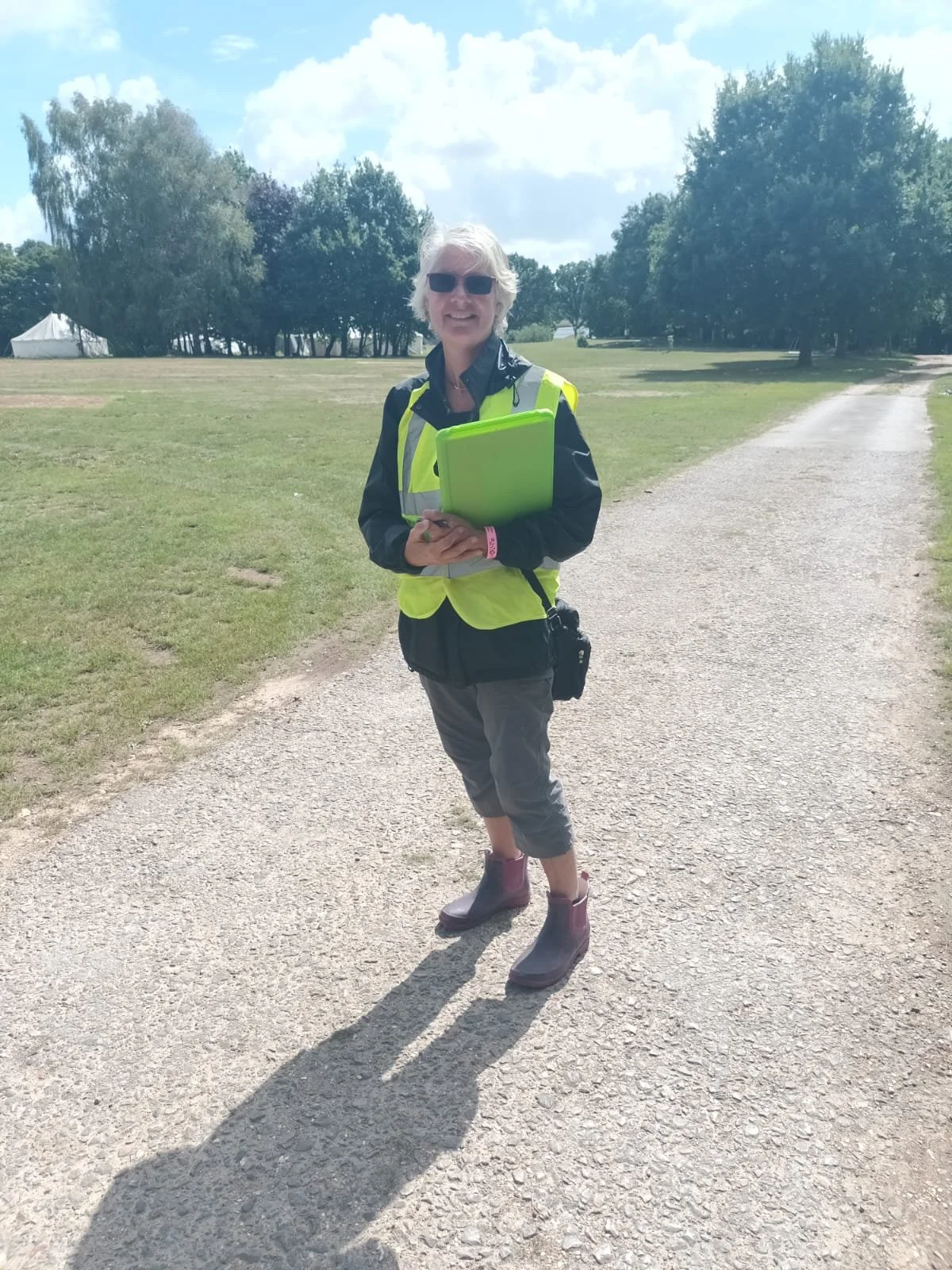 A woman standing on a gravel path in a park, wearing a yellow safety vest, holding a green folder, with trees and a tent in the background.