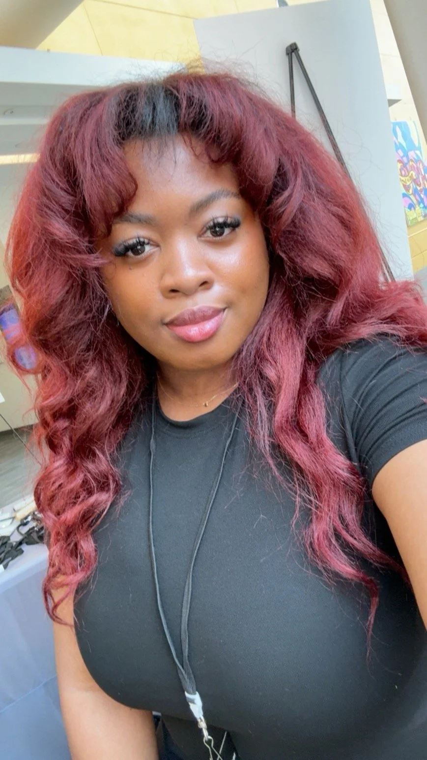 Portrait of a woman with wavy, burgundy hair taking a selfie indoors, wearing a black shirt and a lanyard with an ID badge.