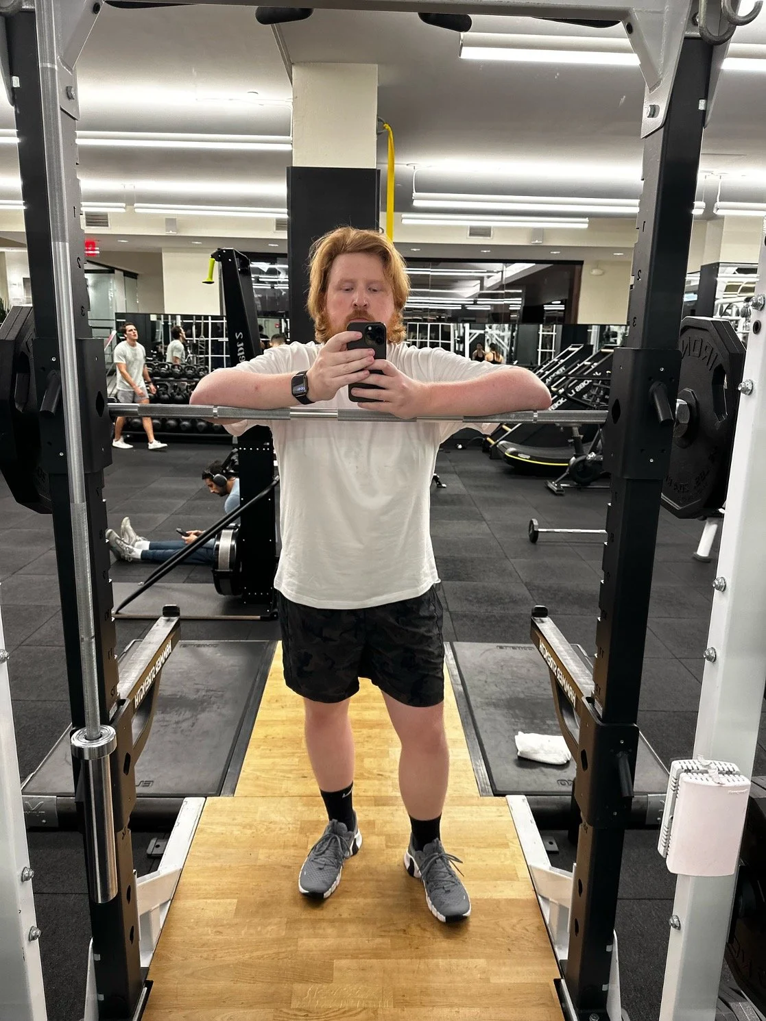 A man with red hair and a beard taking a mirror selfie at the gym. He is wearing a white t-shirt, black camouflage shorts, grey sneakers, and black socks. The gym has various exercise equipment and other people working out in the background.