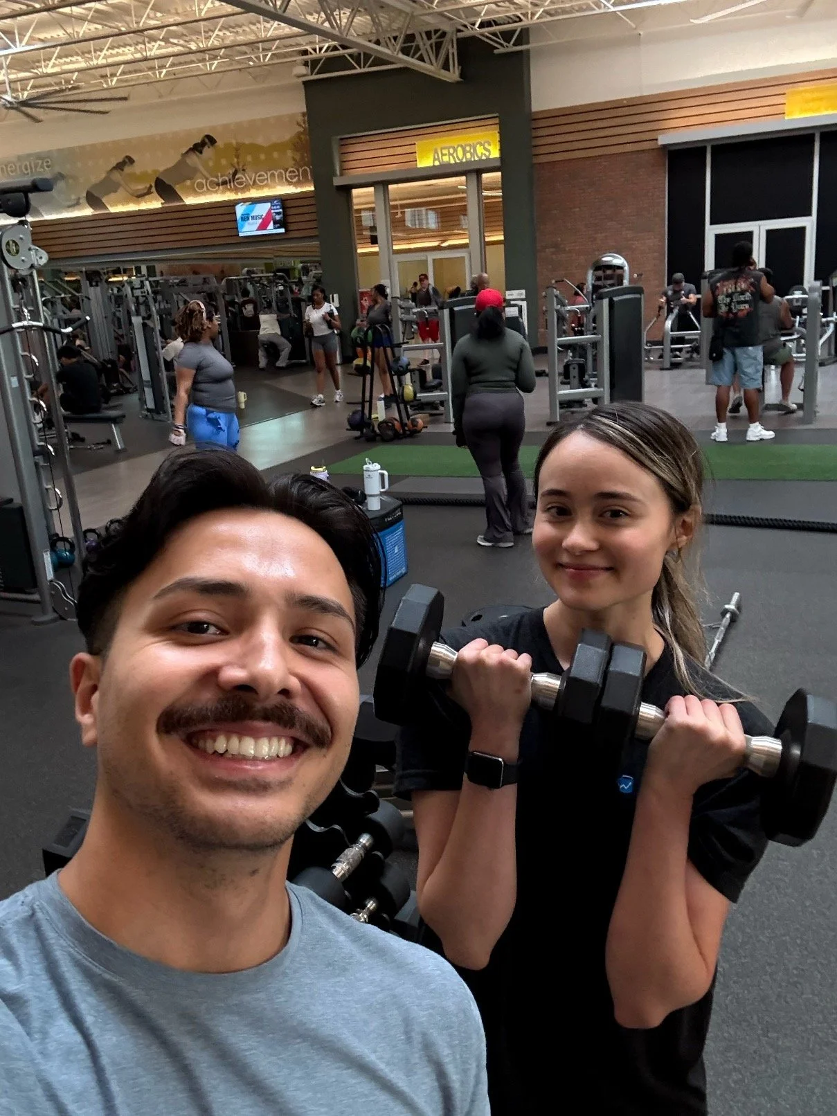 A smiling man and a woman holding a dumbbell at a gym with other people working out in the background.