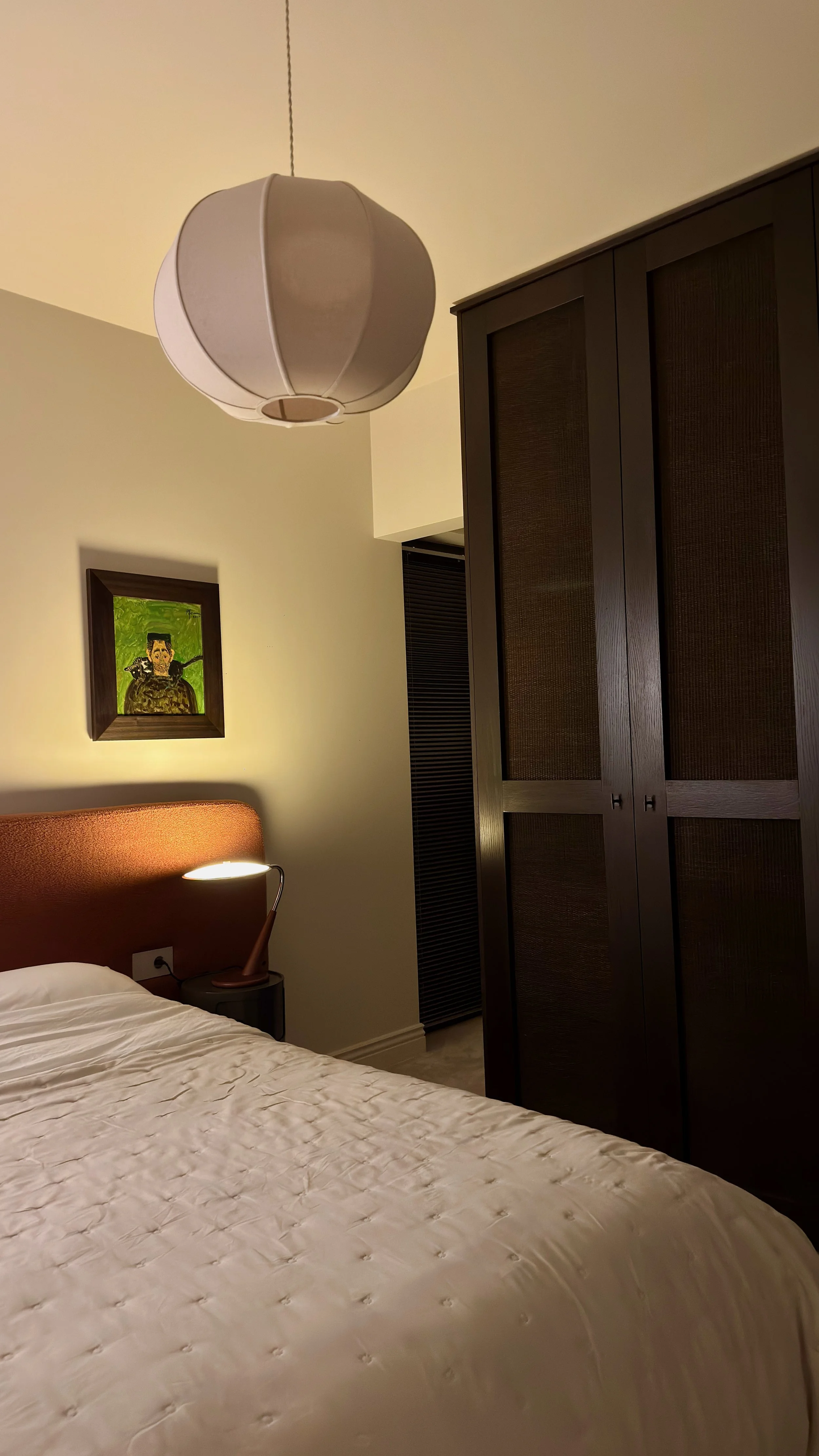 A bedroom with a bed, a bedside table, a wall-mounted lamp, a framed picture, a wardrobe, and a ceiling light fixture.
