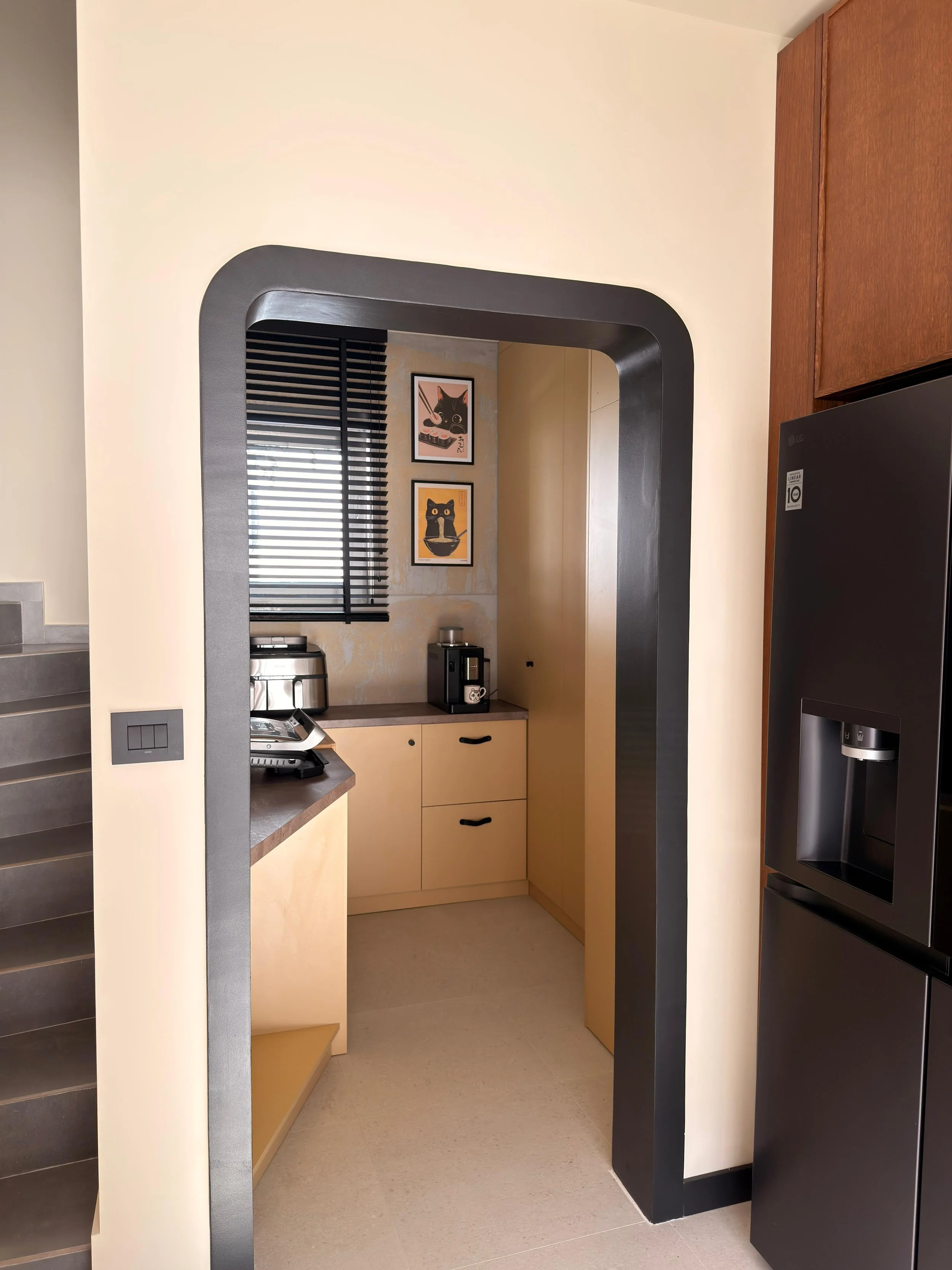 Kitchen with beige walls, black window blinds, a small counter with a toaster and coffee maker, framed cat artwork on the wall, and a black refrigerator.