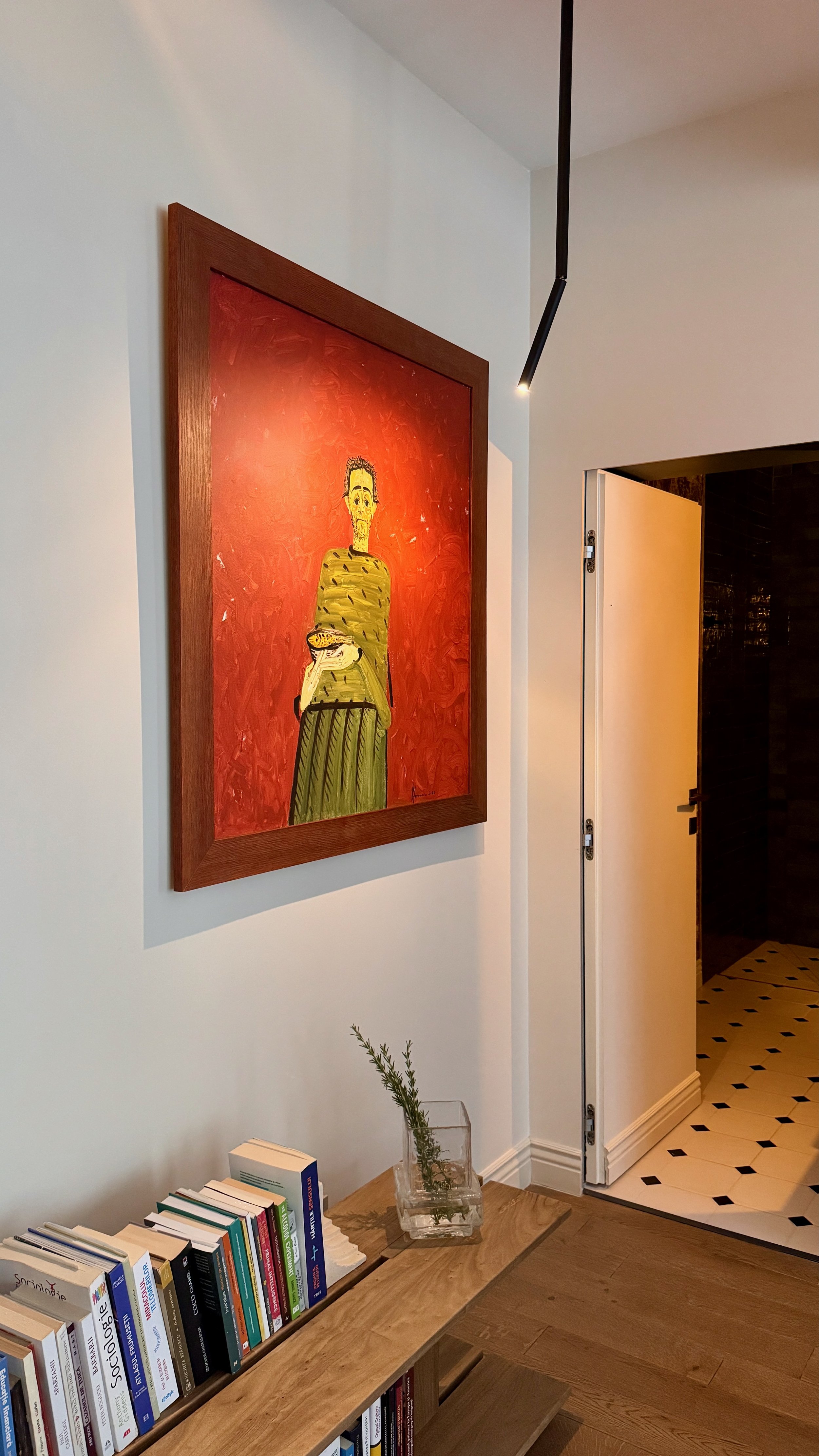 Interior view of a room showing a framed painting of a person holding a yellow fish on a red background, a small table with books, and a glass vase with a sprig of greenery, near an open door.
