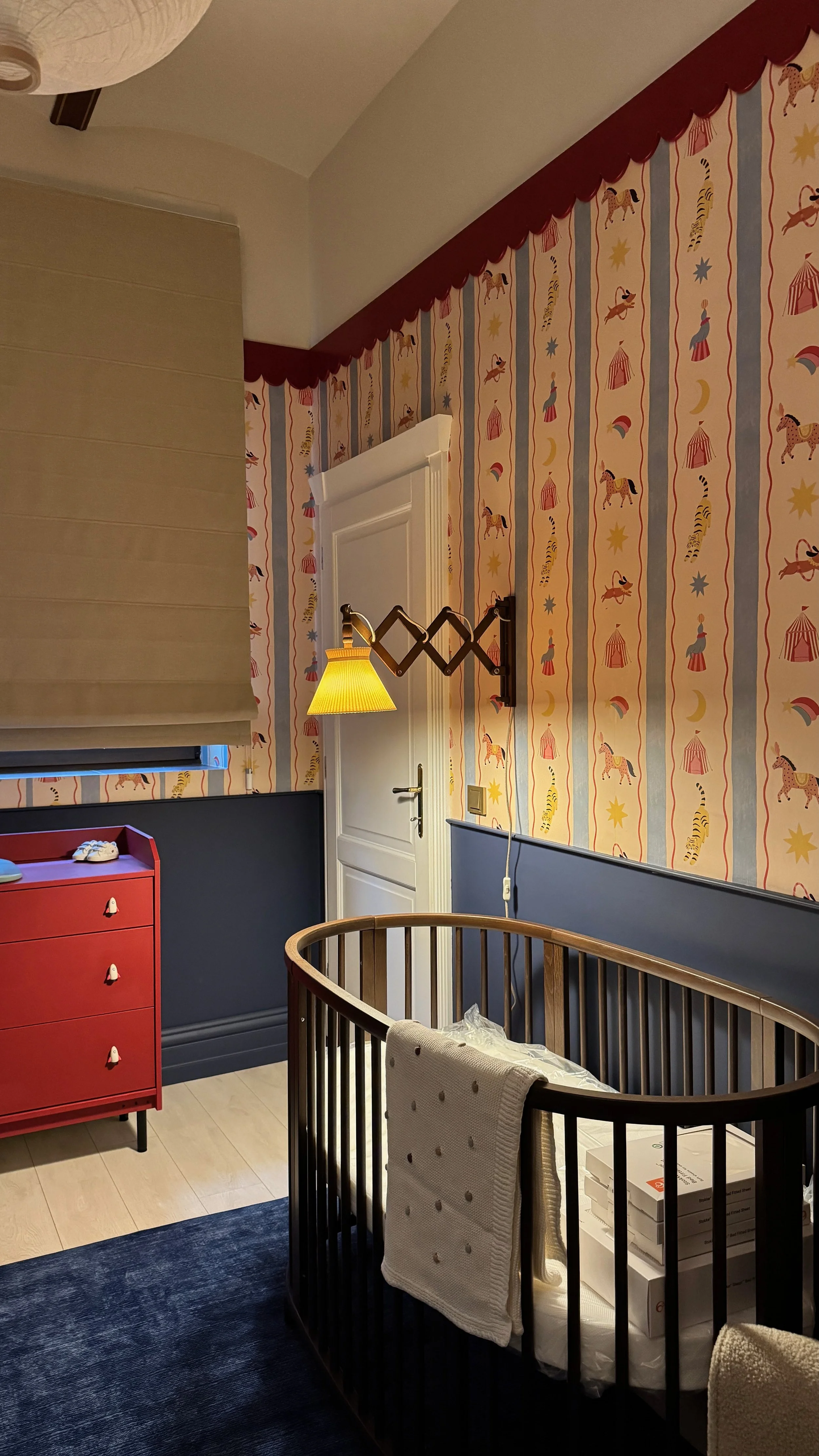 A nursery room with wallpaper featuring circus animals and tents, a white door, a wall-mounted lamp, a red dresser with toys, a black crib with packages inside, and a beige towel hanging over the crib's side.