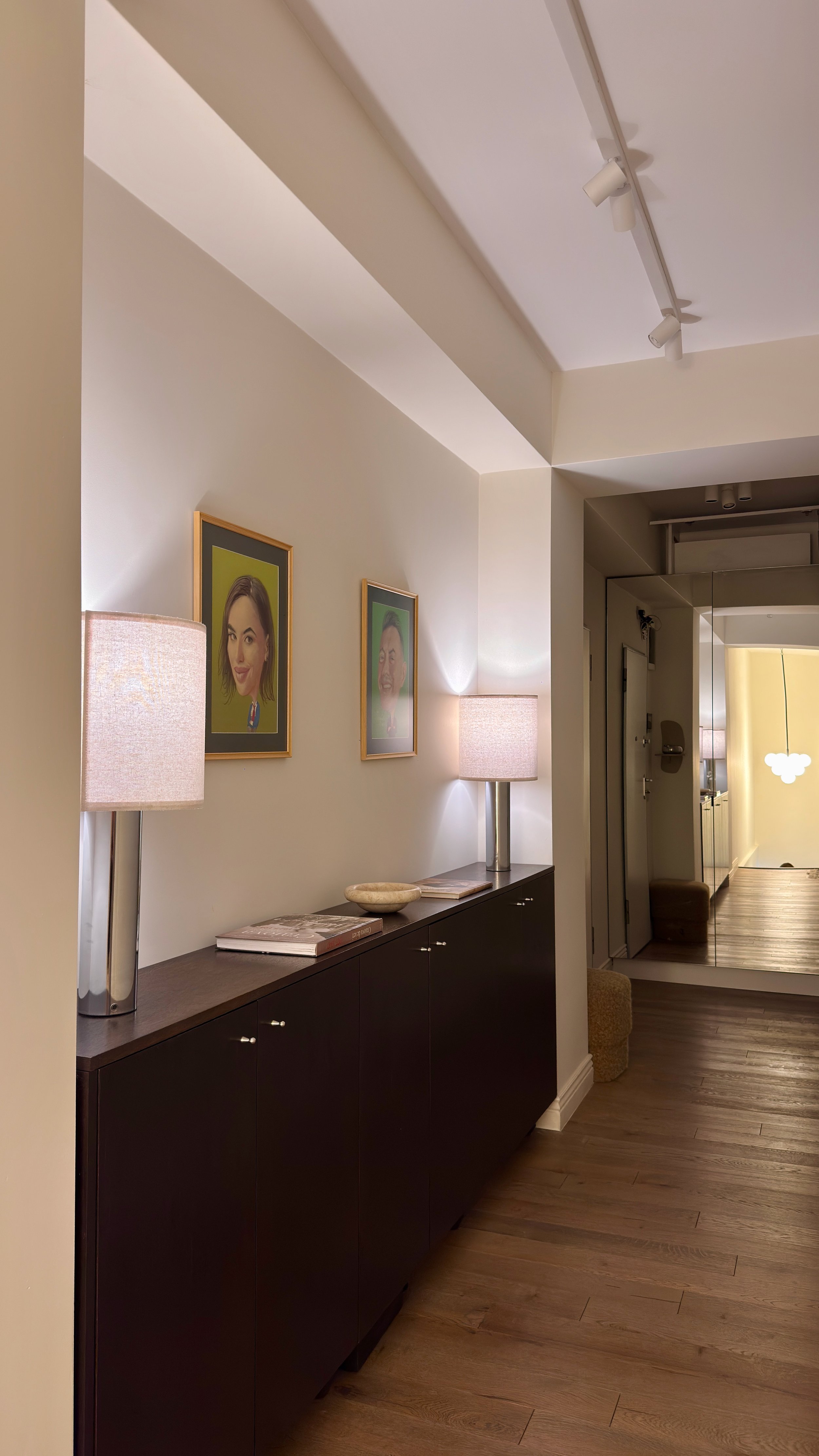 Photo of a hallway with a black sideboard, two framed caricature portraits on the wall, two table lamps, and a mirror reflecting part of a room. Wooden floor and a small stool are visible.