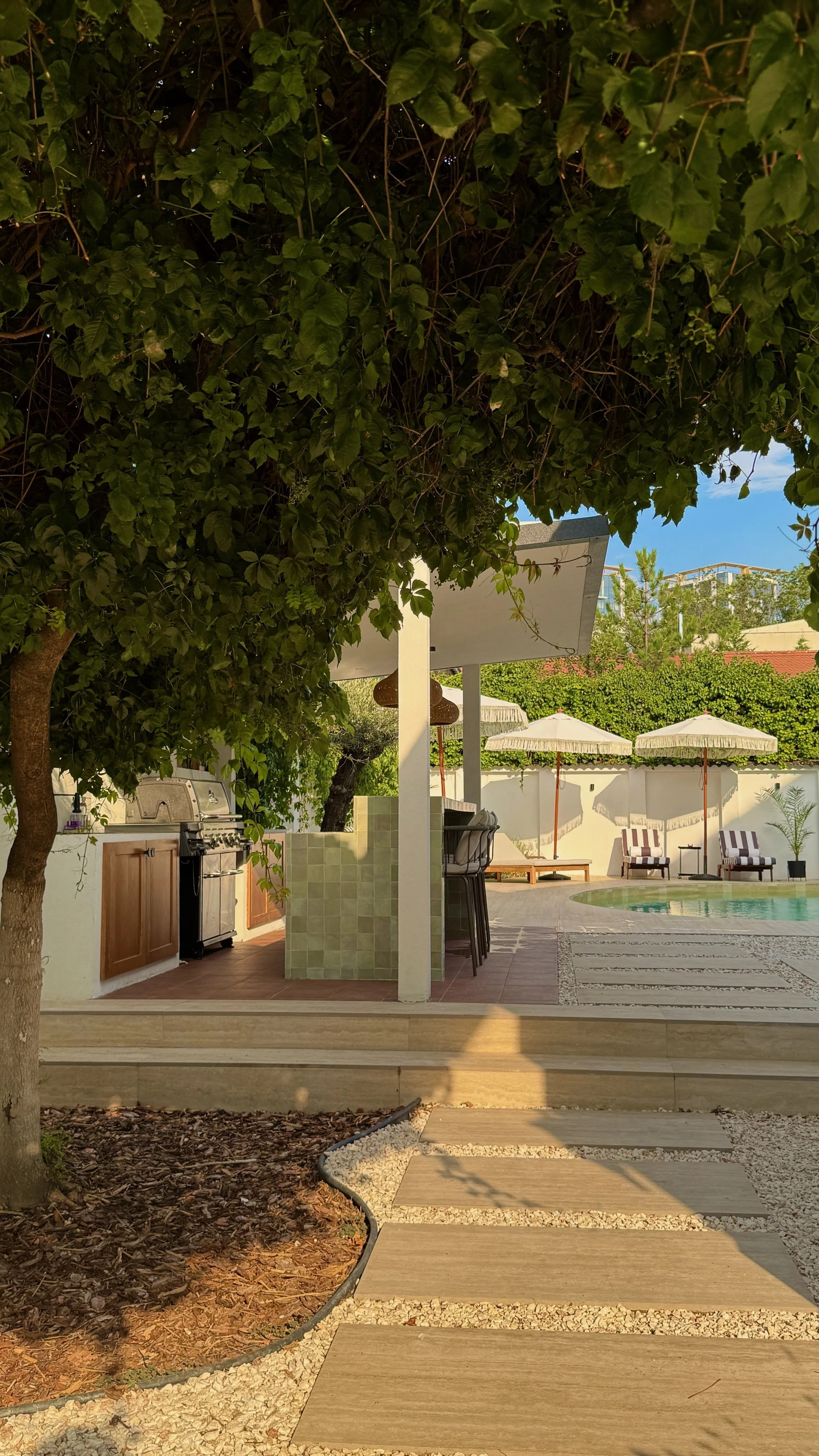 A backyard poolside area with a shaded bar or seating area under a roof with a barbeque grill on the side. There are white umbrellas and lounge chairs next to the pool, and trees and greenery surround the area.